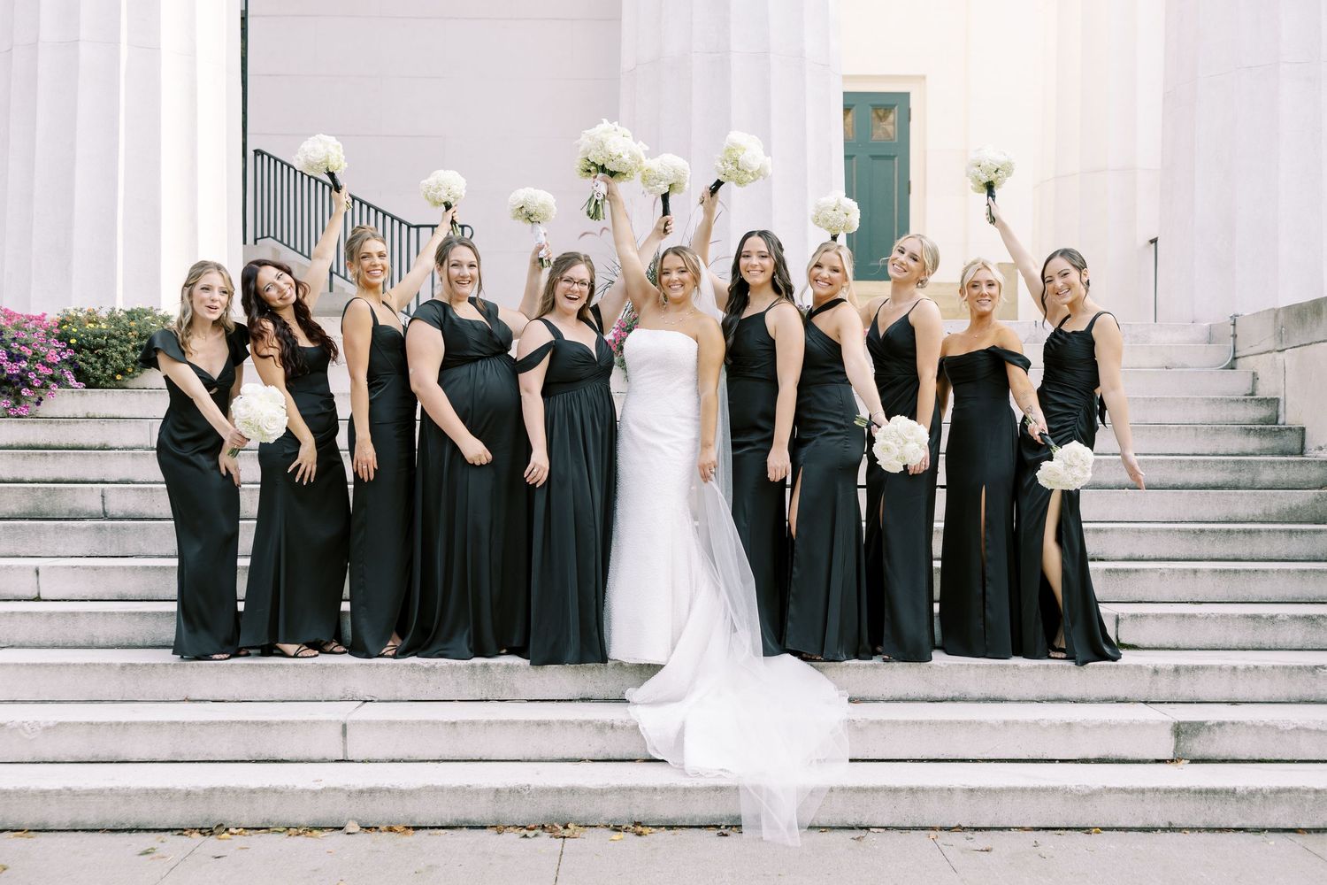 Wedding party in black dresses and white gown pose together on stone steps for formal group photos.