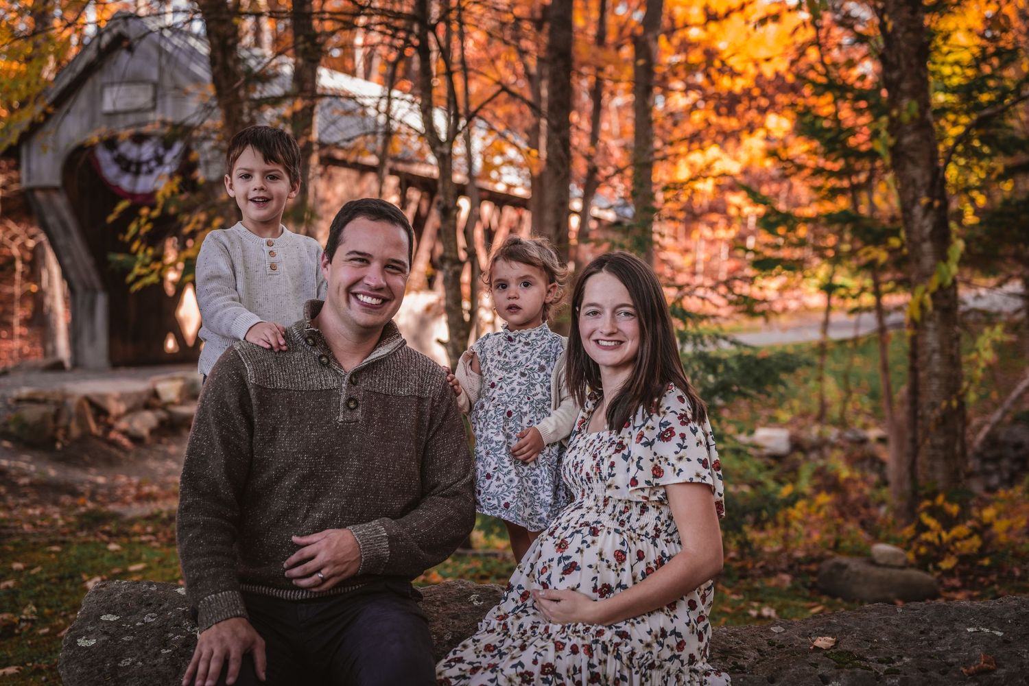 Happy family of four with pregnant mother posing outdoors surrounded by vibrant fall foliage.