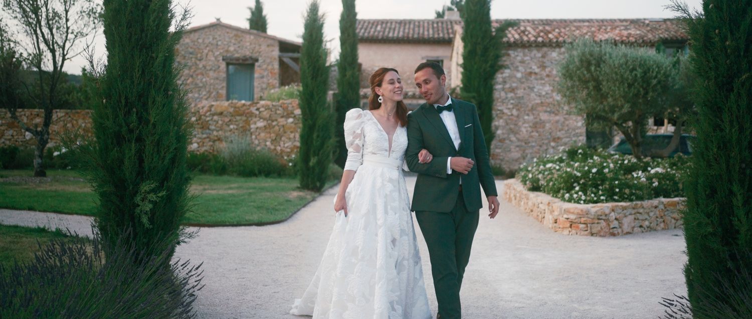 A couple stands together in a romantic outdoor setting with Italian cypress trees and stone buildings.