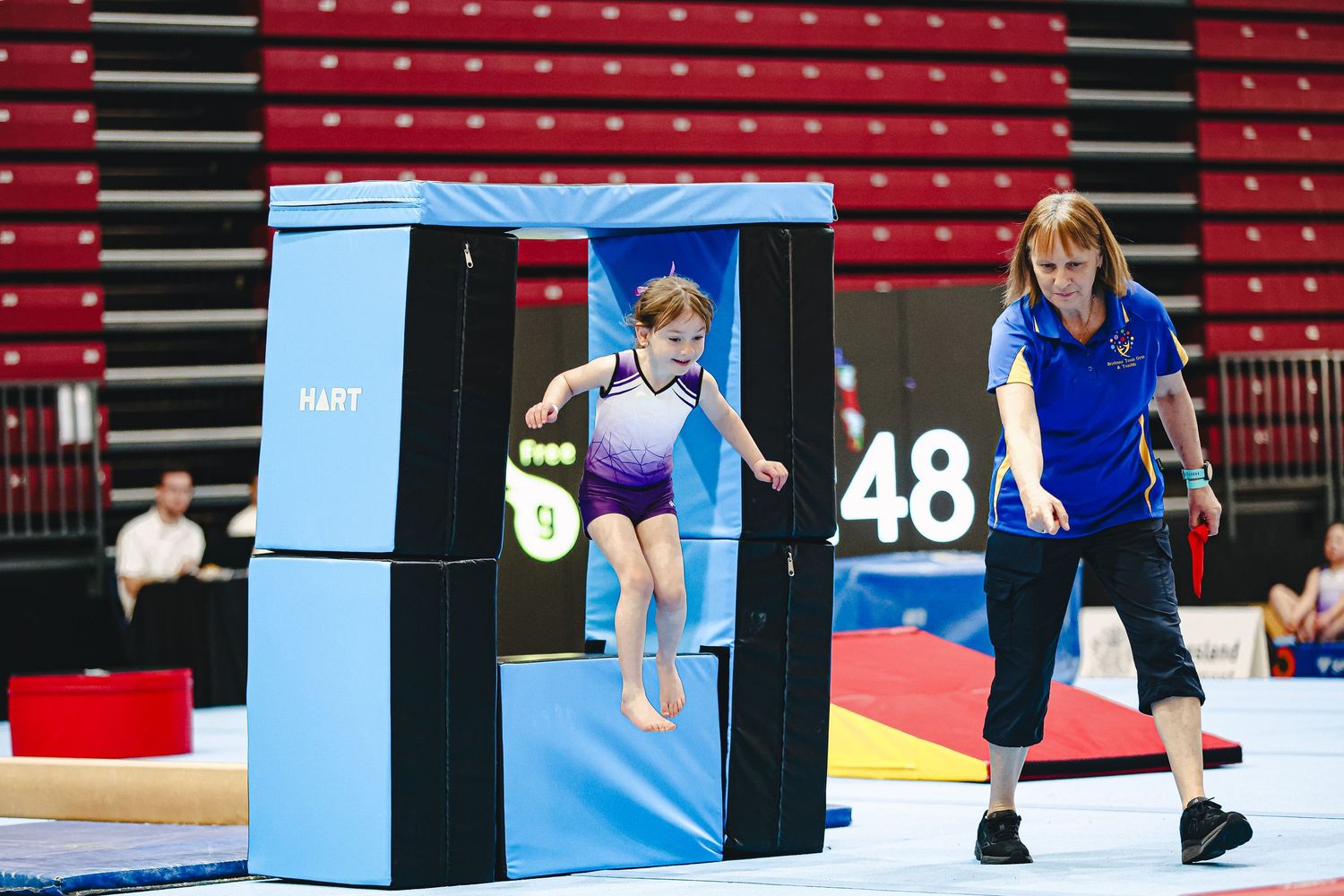 Young gymnast in blue leotard practices jumps on gymnastics equipment with instructor nearby in red stadium.