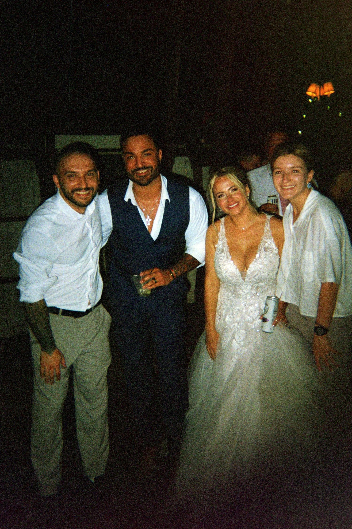 A group poses together at a wedding celebration with the bride in a white lace gown and guests in formal attire.