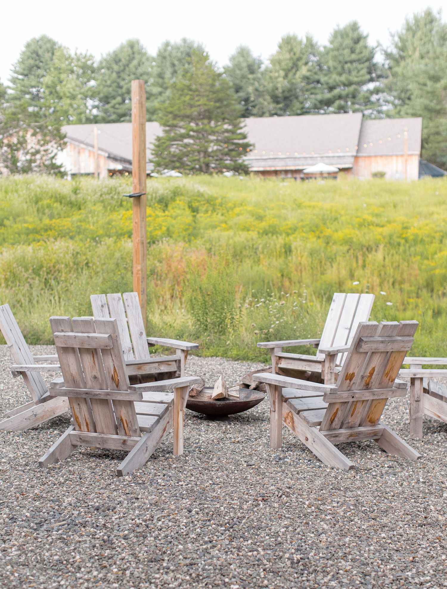 Rustic Adirondack chairs arranged around a fire pit with wildflower meadow and building in background.