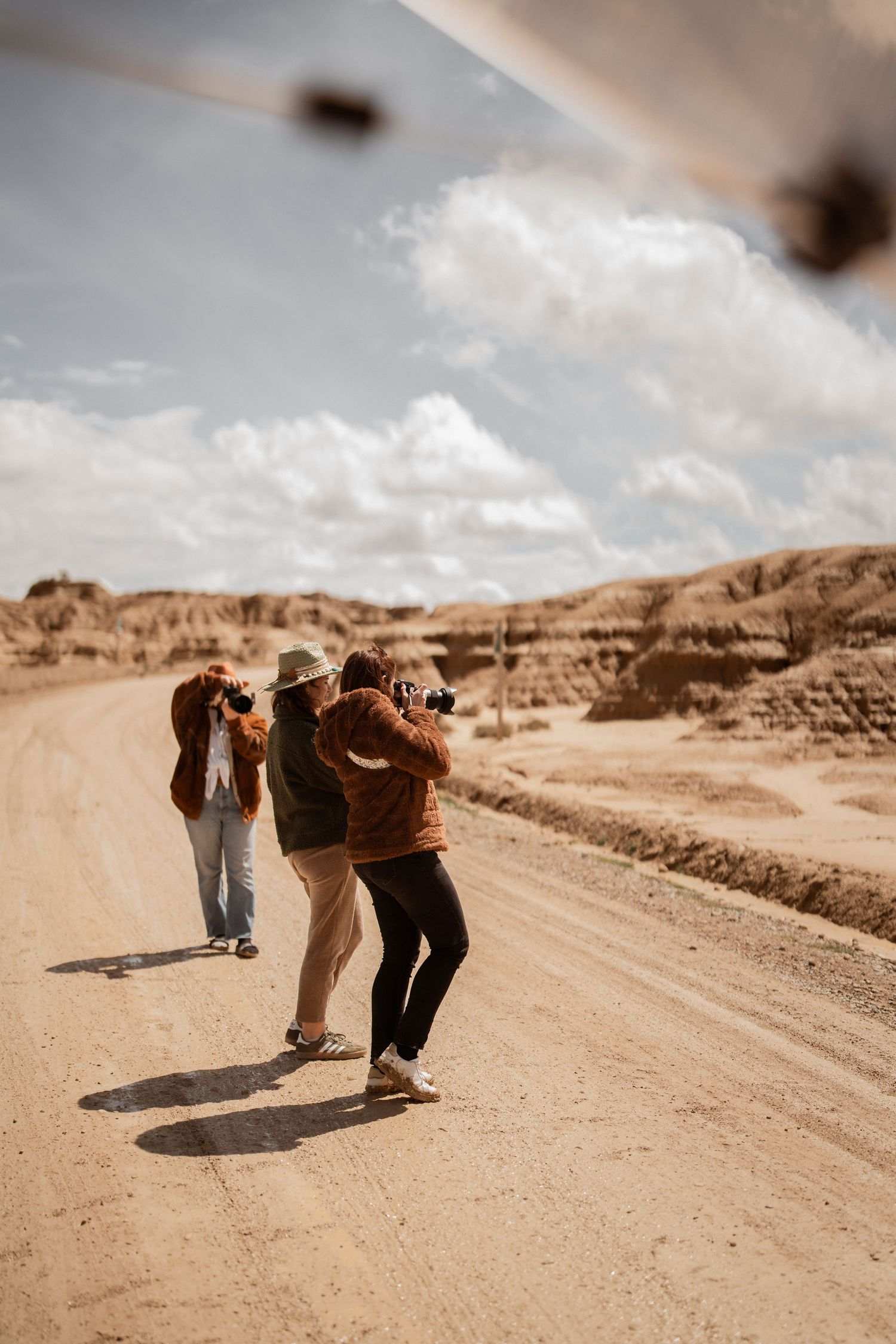 Photographers with cameras capturing shots on a dusty desert road surrounded by rugged rock formations under cloudy skies.
