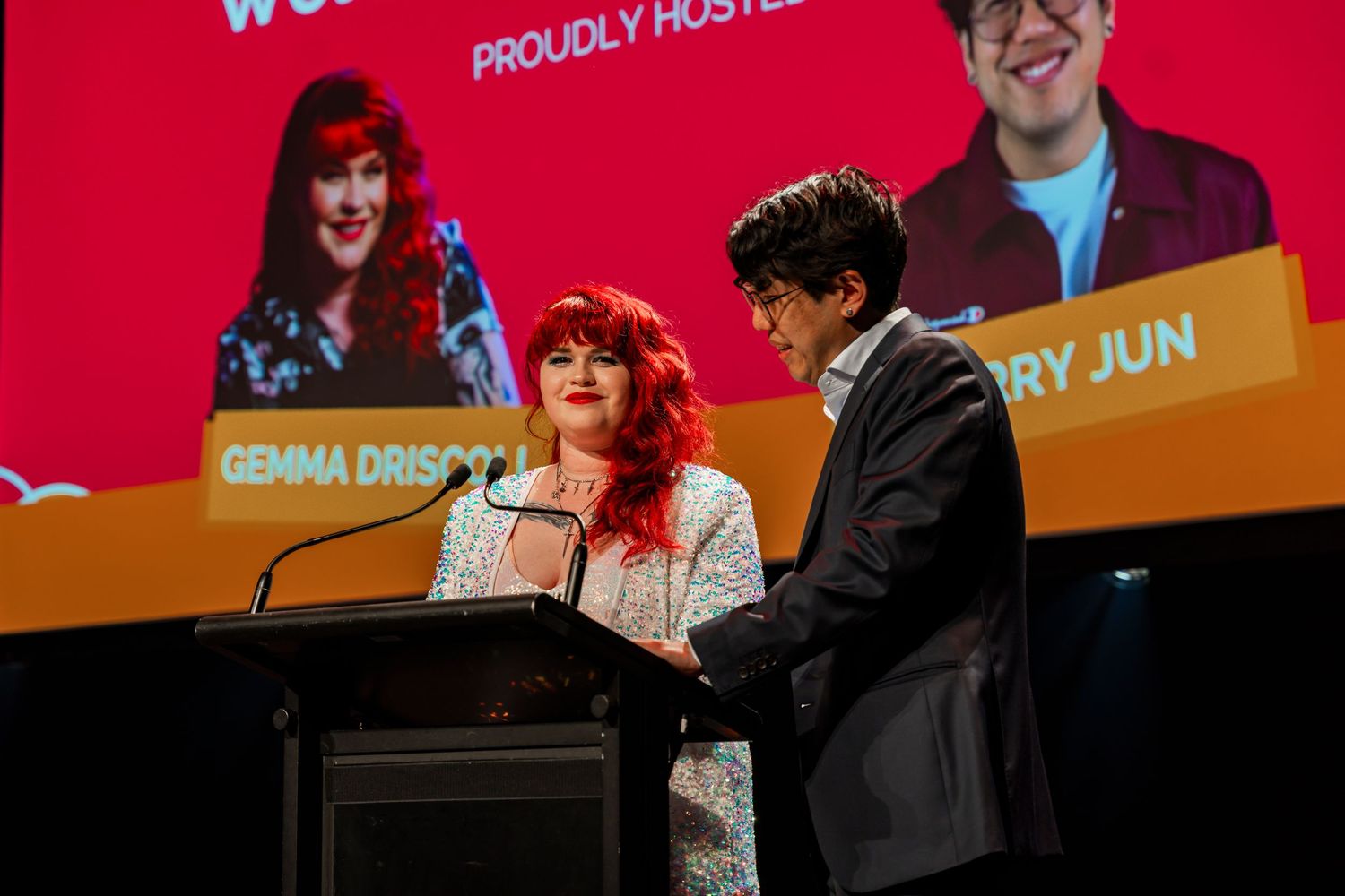 Two people standing at a podium with a bright red and orange backdrop during an event presentation.