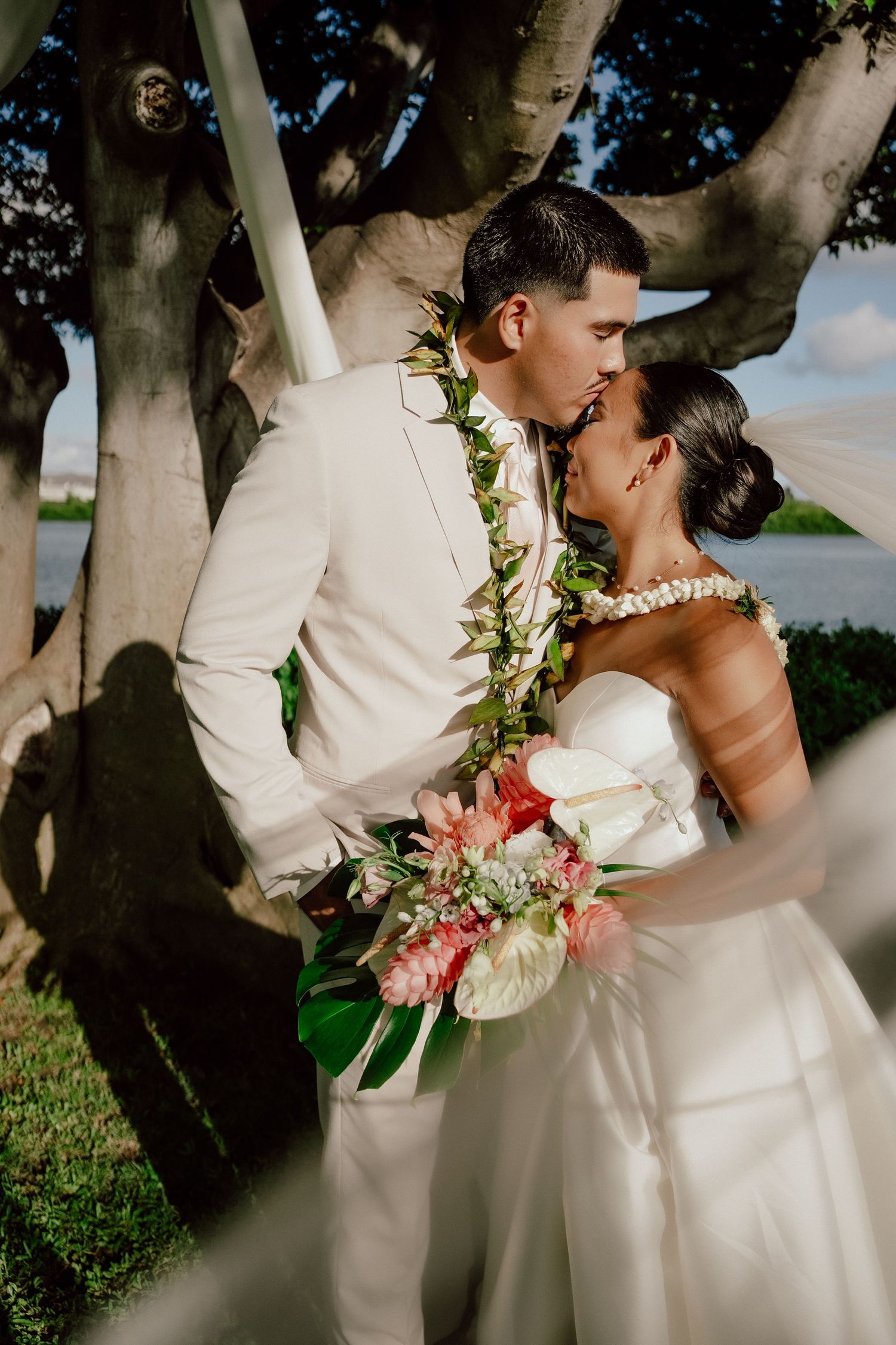 Two people in white wedding attire embrace under trees in a romantic outdoor wedding photoshoot.