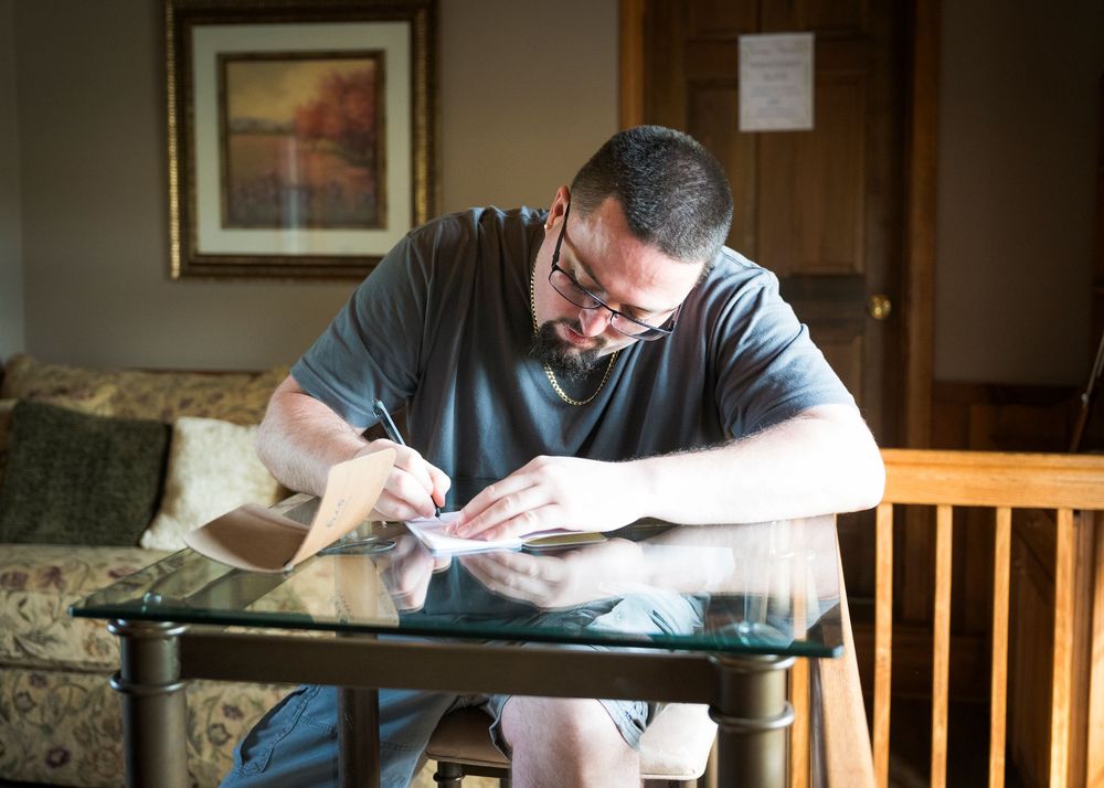 Person writing at a glass table in casual attire near a window.
