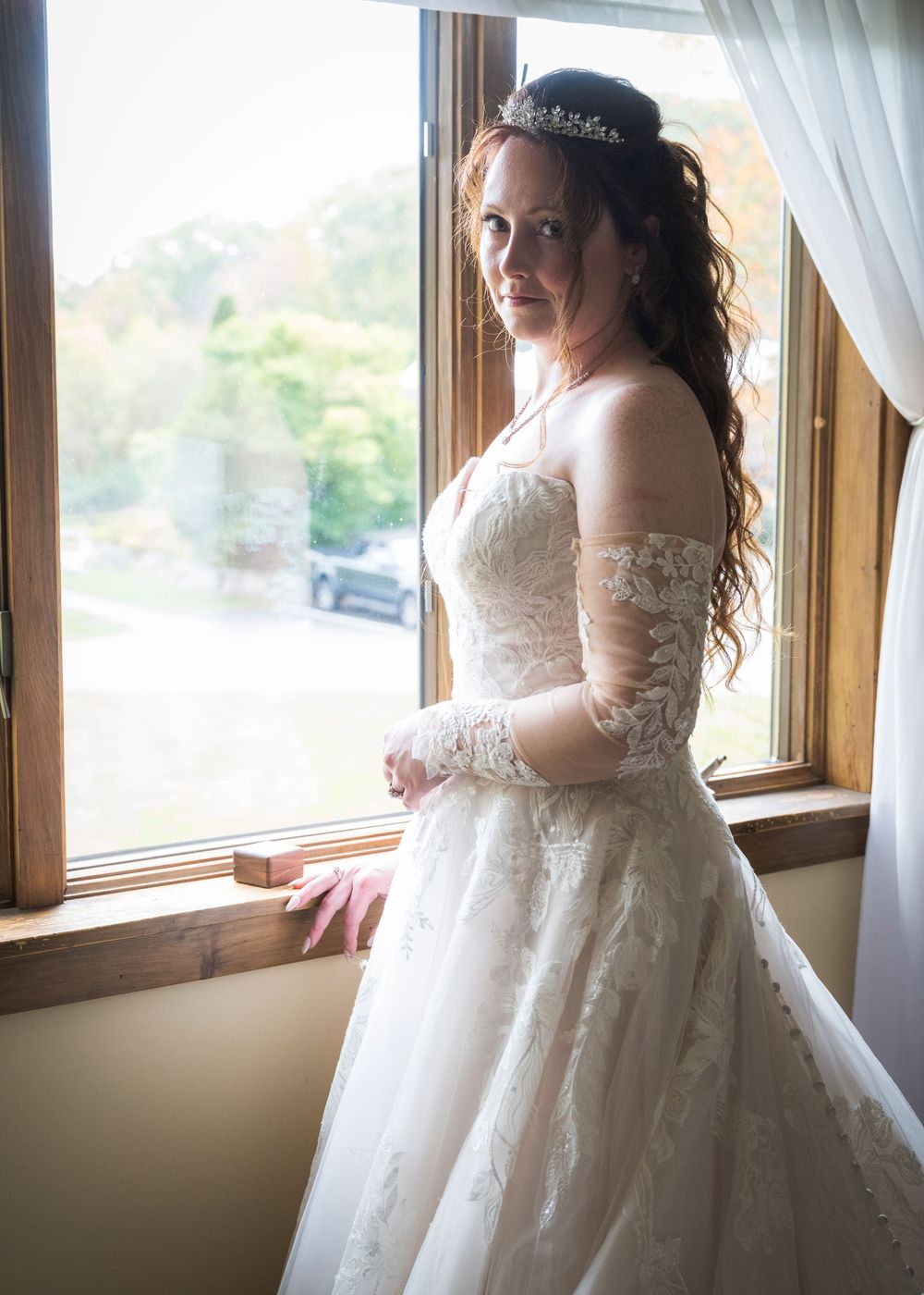 A wedding dress with intricate lace details and long sleeves is showcased by a window with natural light.