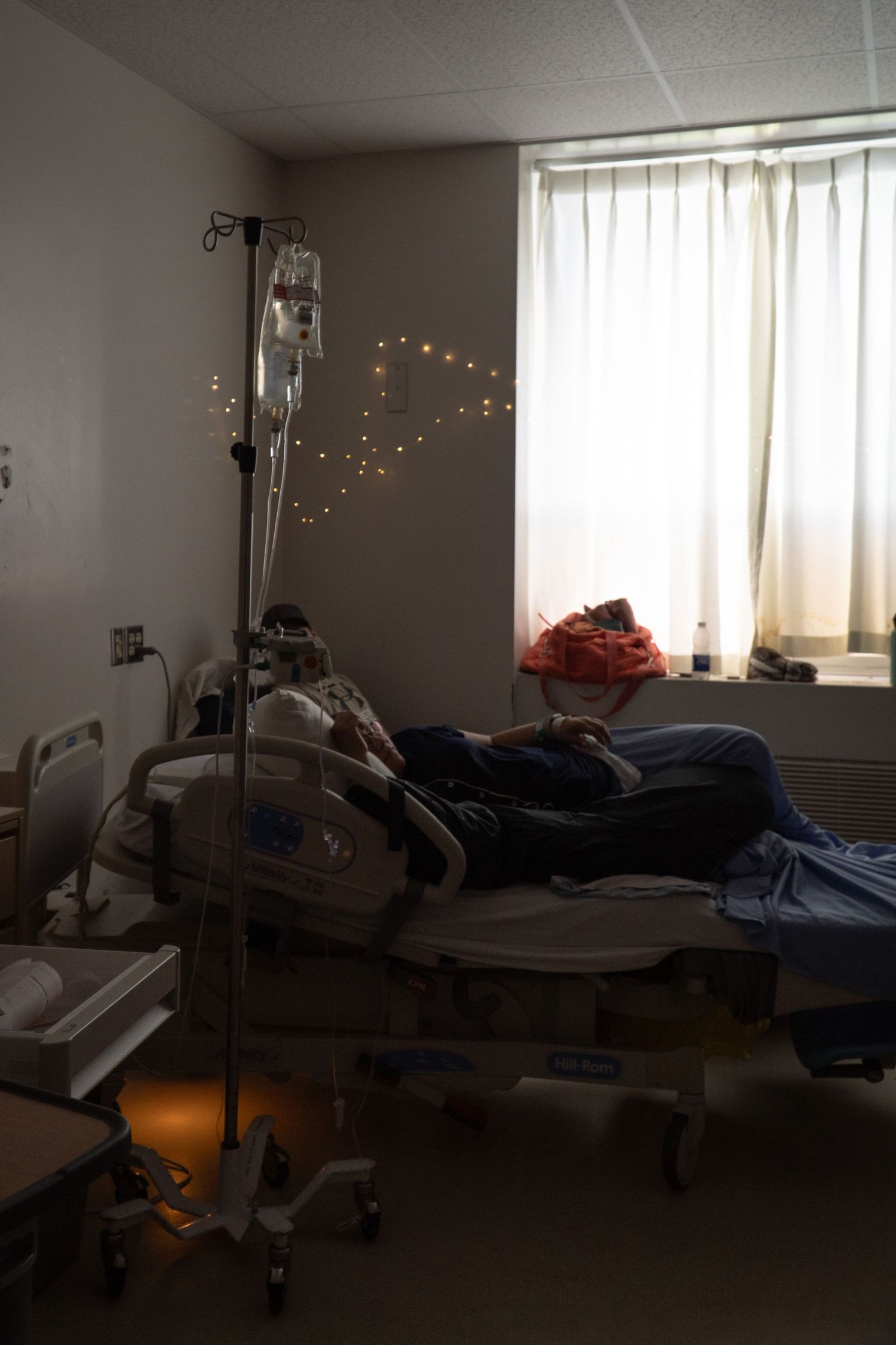 Pregnant woman resting in a dimly lit hospital room with IV drip and fairy lights by window.