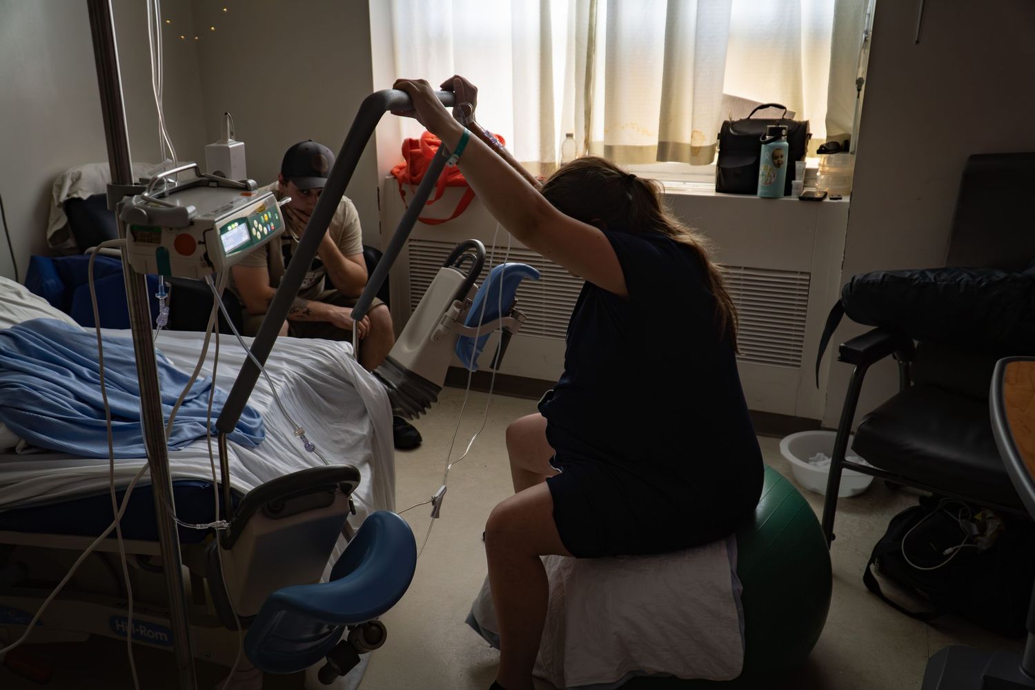 Woman in labor using a peanut ball for support while partner assists in a hospital room.
