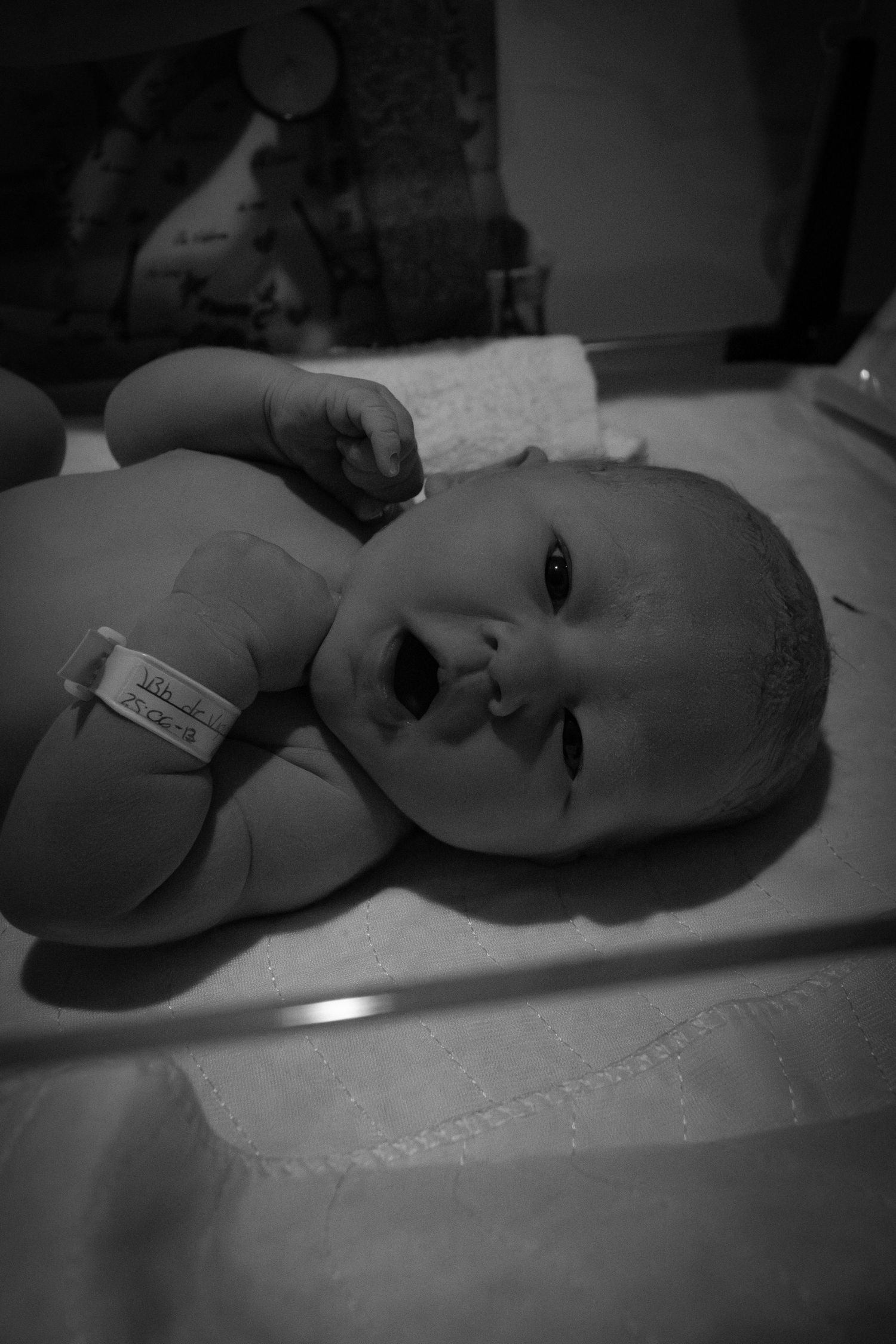 Black and white close-up portrait of a newborn baby with hospital ID bracelet on exam table.