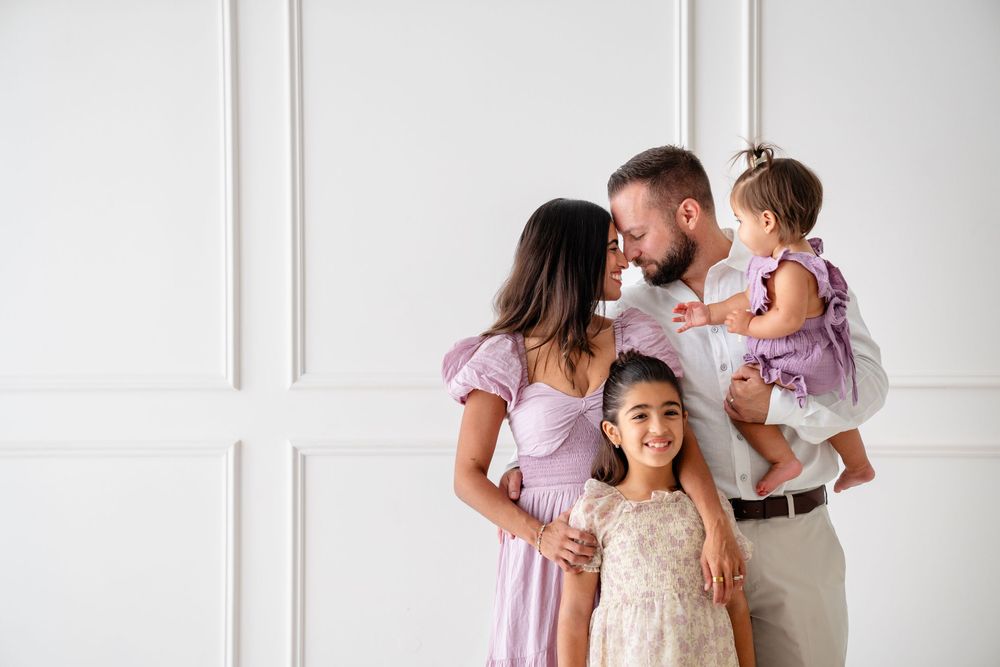 Family wearing coordinated pink and white outfits poses together against a white wall with decorative molding.