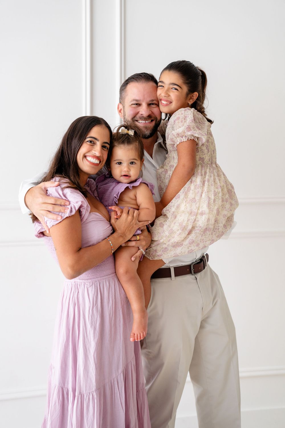 Happy family wearing pastel outfits embraces together against white wall during portrait session.