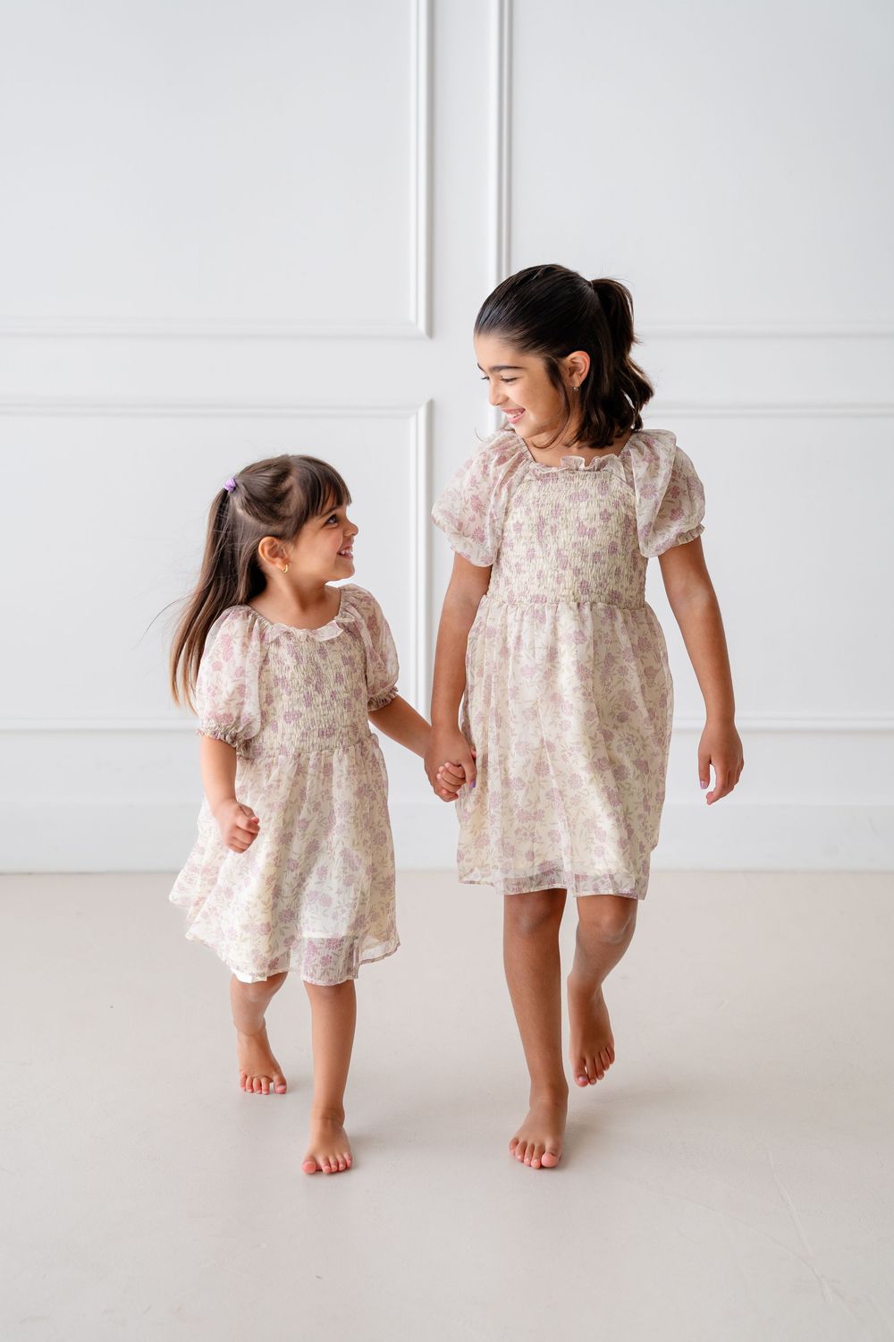 Two people in matching floral dresses hold hands and walk together in a bright studio setting.