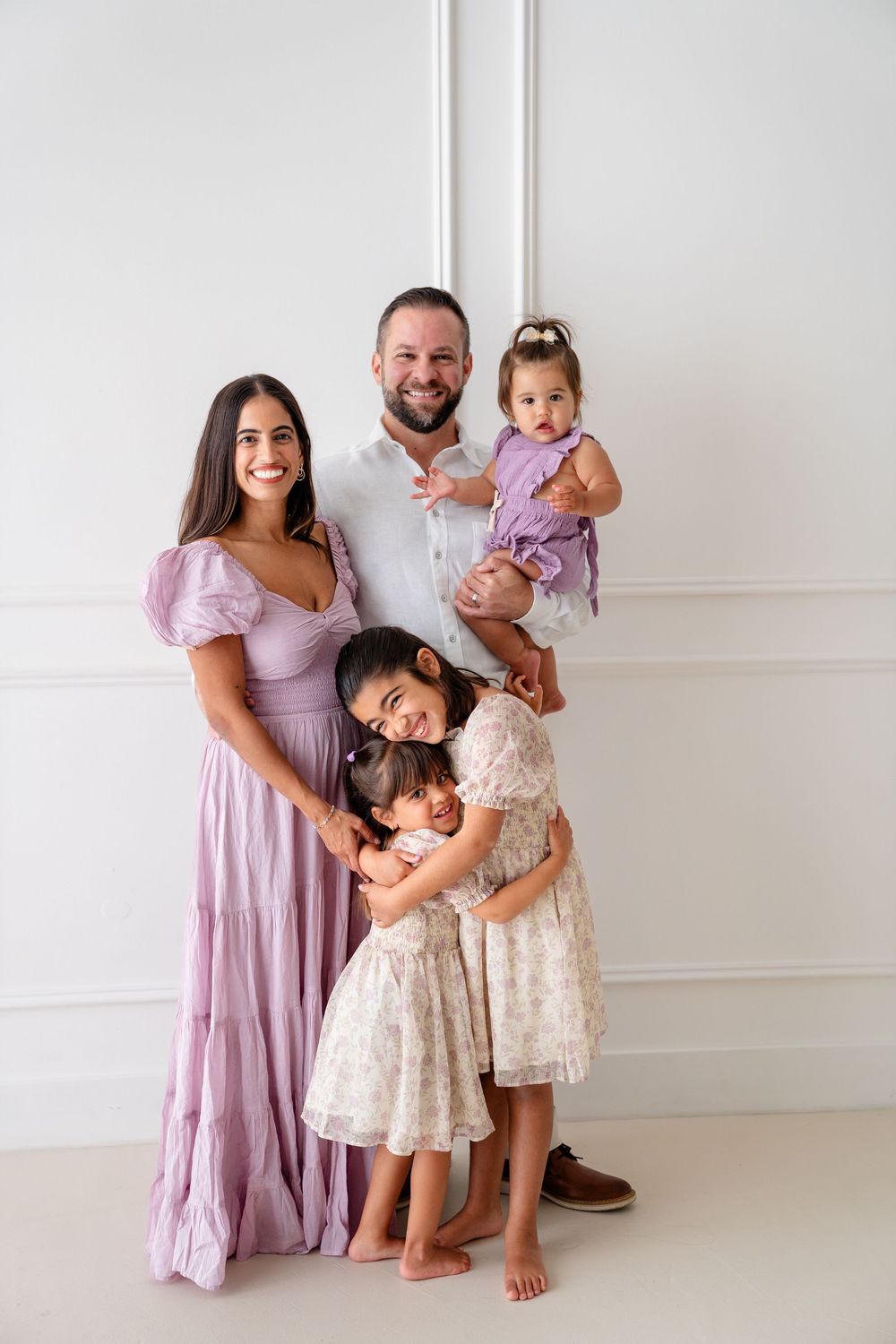 A joyful family moment with parents and children wearing coordinated lavender and white outfits against a white wall.
