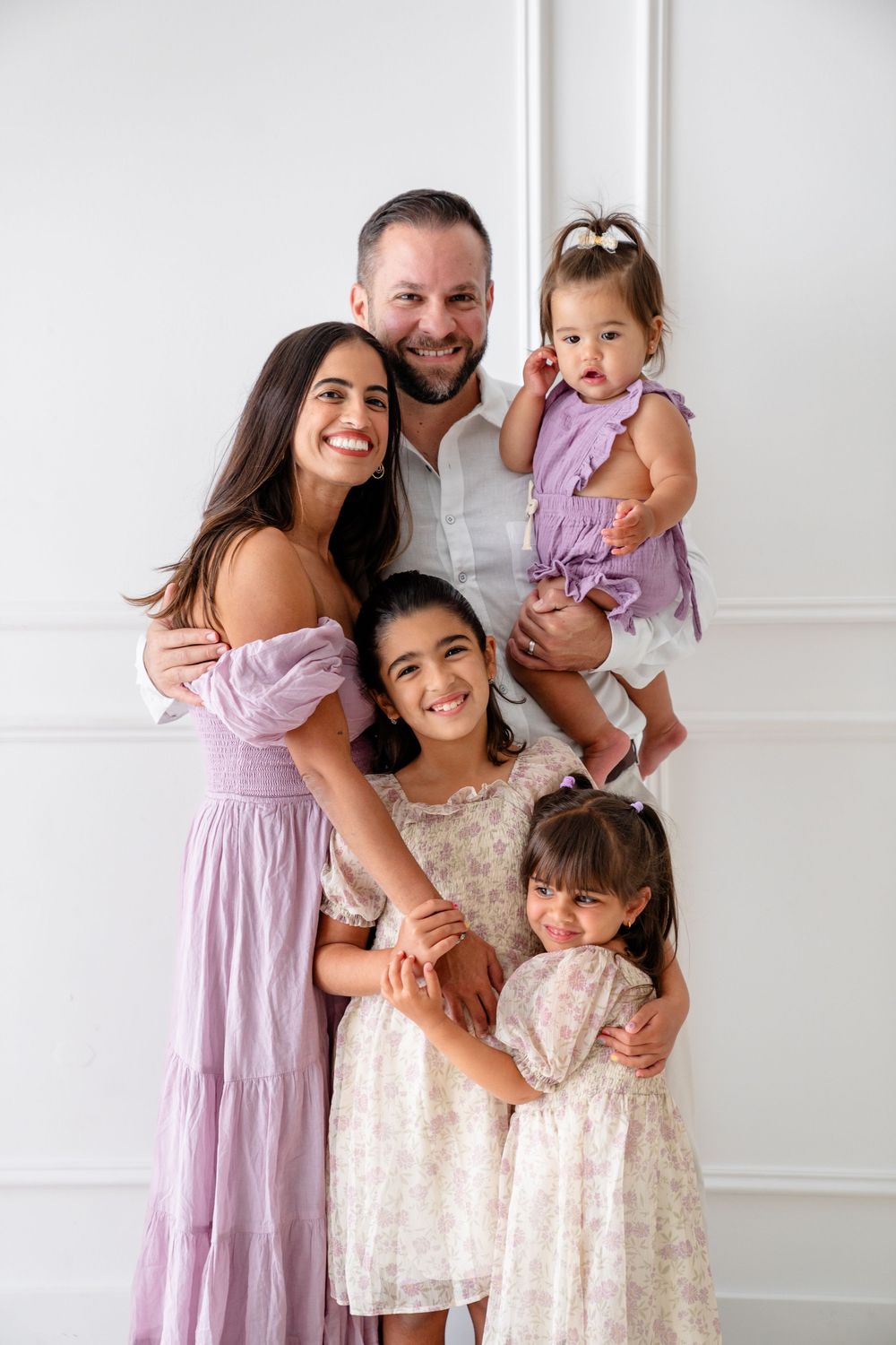 Family portrait featuring coordinated purple and cream dresses against a white wall with decorative molding.