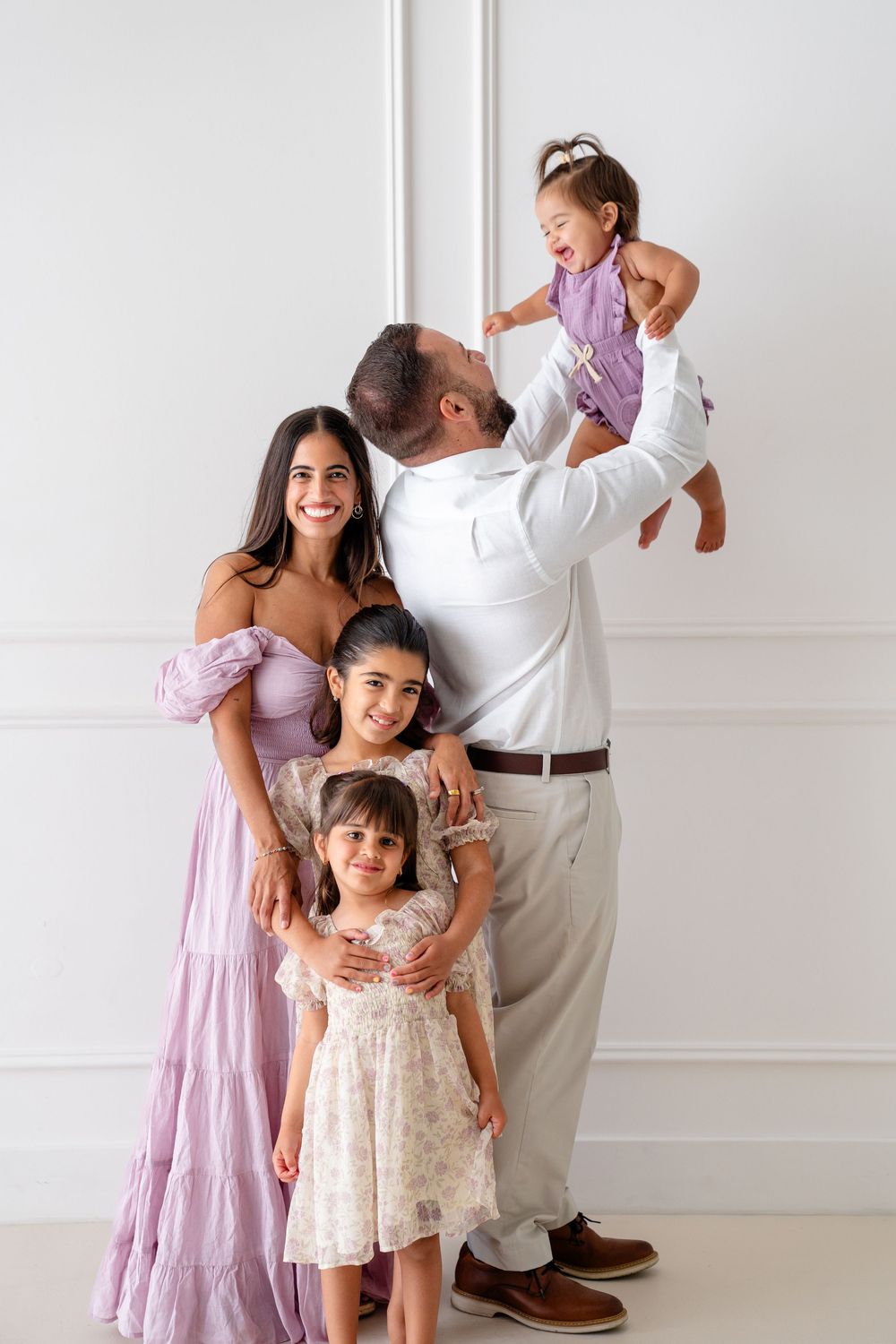 Family enjoying playful moments together in a bright white studio wearing lilac and cream outfits.