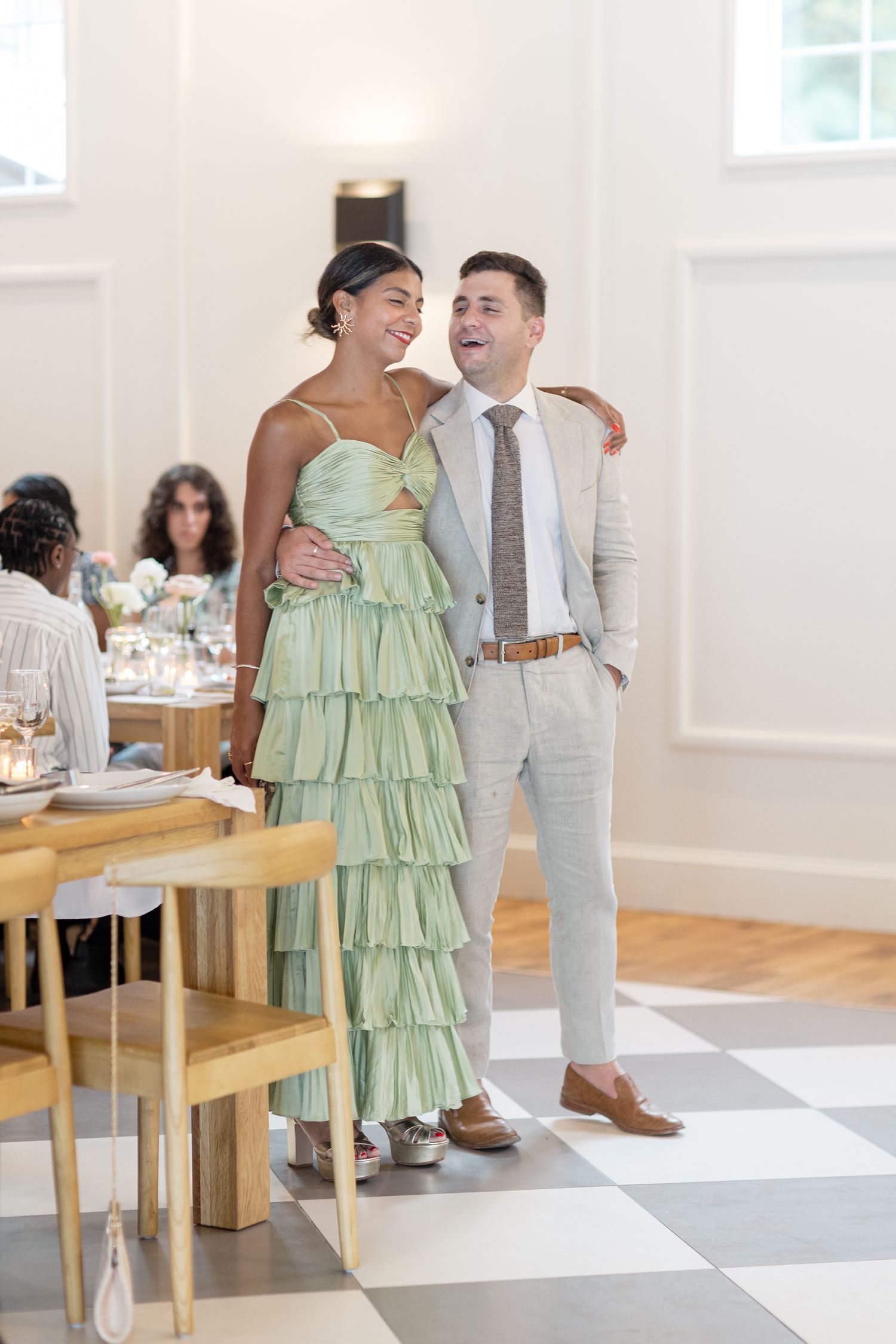 Guests share candid laughing moments while sitting in wooden chairs at a wedding reception celebration.