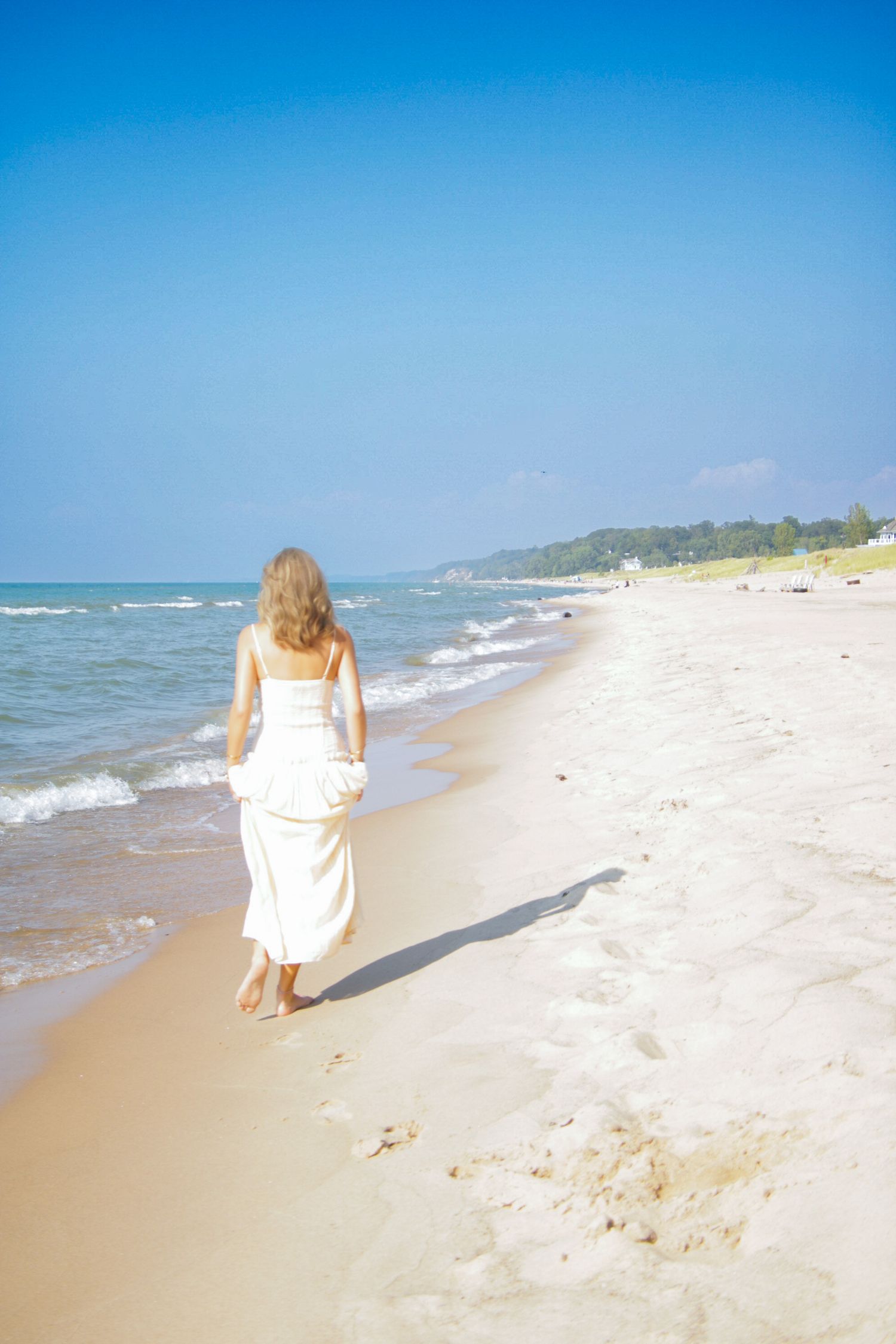 Person in white dress walks along pristine beach shoreline with blue ocean waves and clear sky.
