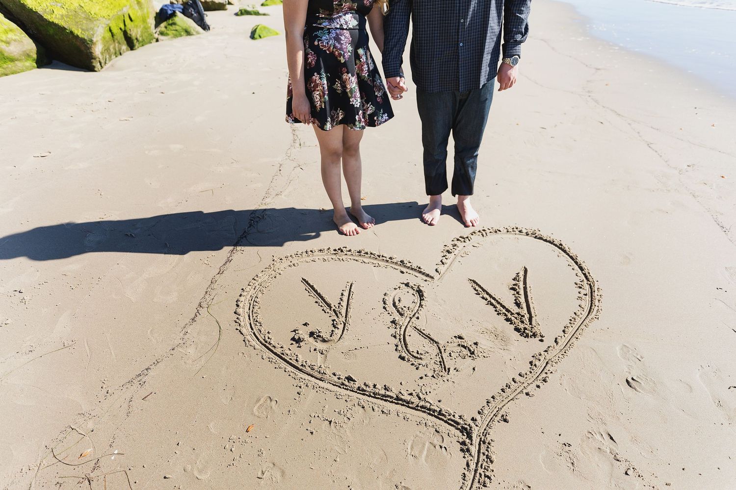 Heart shape with initials drawn in beach sand with a couple's feet standing nearby at the shoreline.