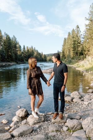 A couple explores a rocky riverbank with pine trees in the background.