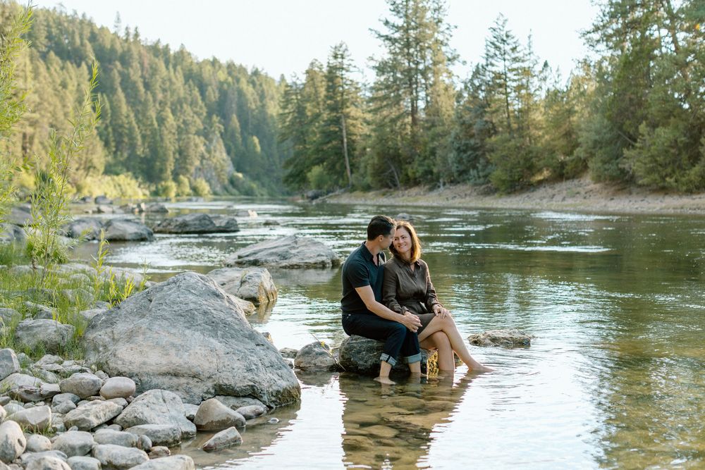Couple enjoying a romantic photoshoot by a scenic river surrounded by evergreen trees and rocky shores.