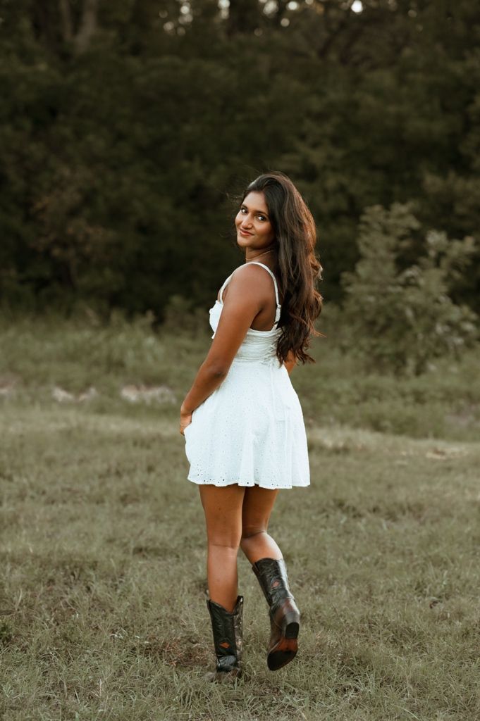 A woman in a white summer dress and cowboy boots poses playfully in a grassy field during golden hour.