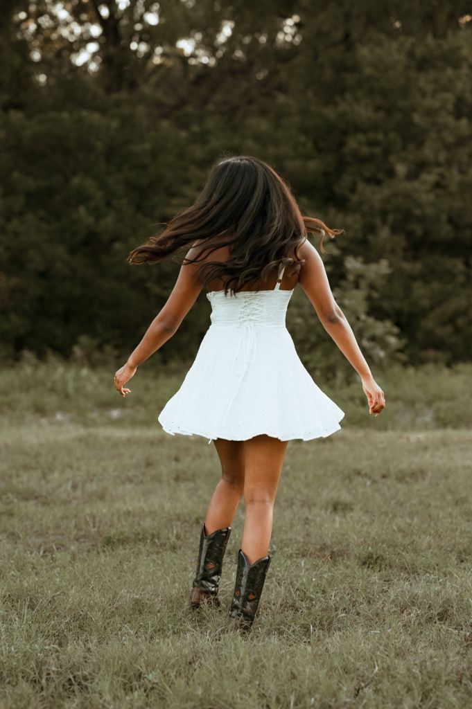 A person in a white dress and black boots spins playfully in a grassy field at sunset.
