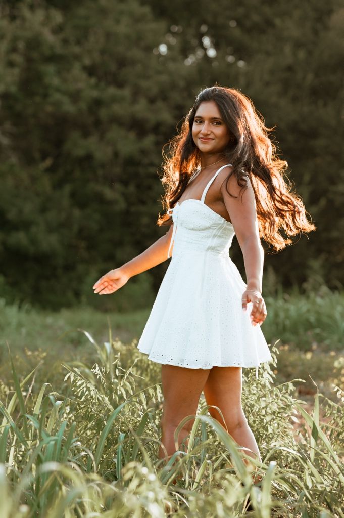A woman in a white summer dress spins joyfully through a sunlit meadow of tall grass at golden hour.