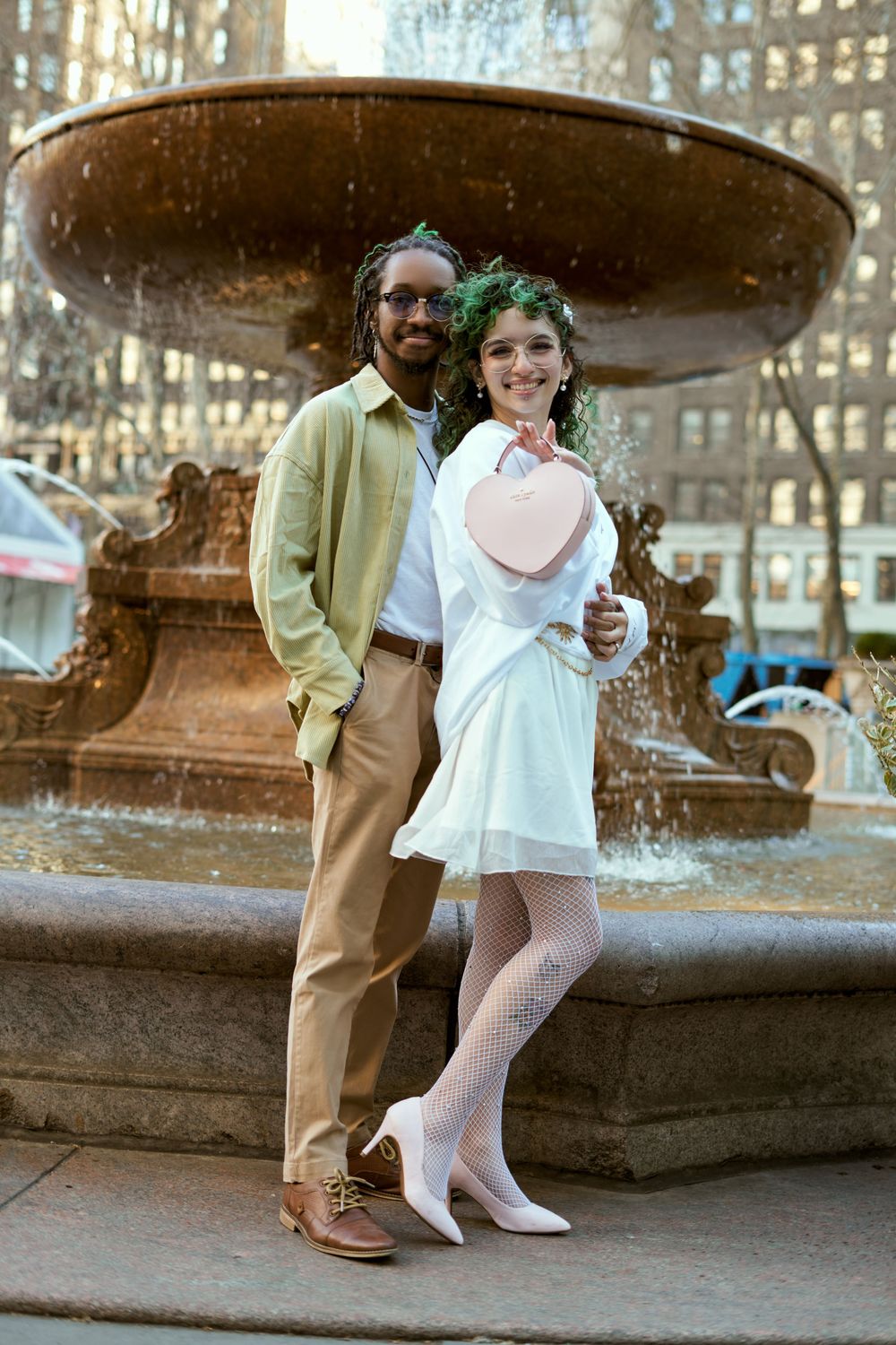 A couple poses together by a large fountain in a romantic outdoor setting with city architecture in the background. Engagement shoot Manhatt