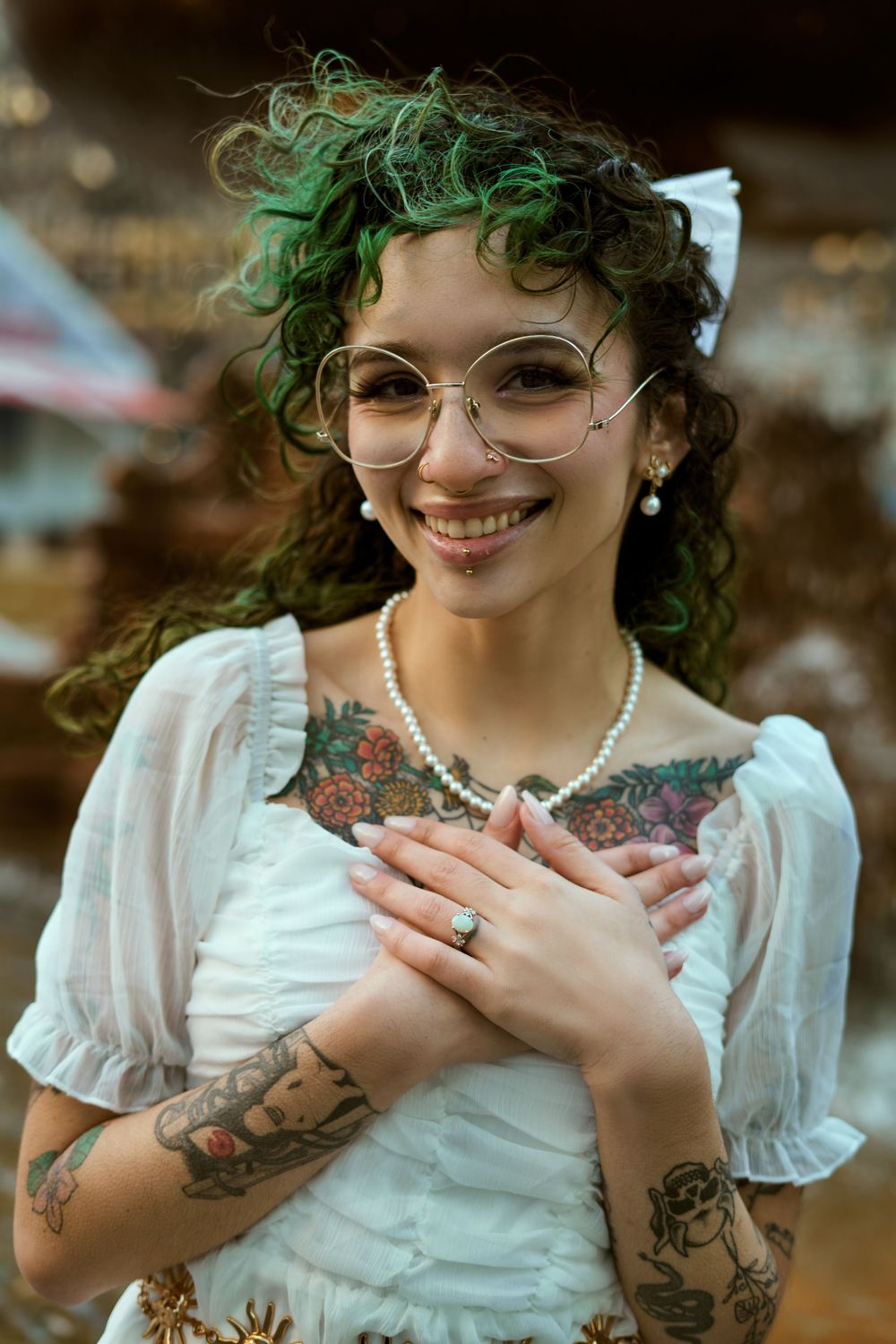 Portrait of an alternative style bride wearing glasses, white dress, green curly hair, pearls and vibrant tattoos on their arms. Engagement