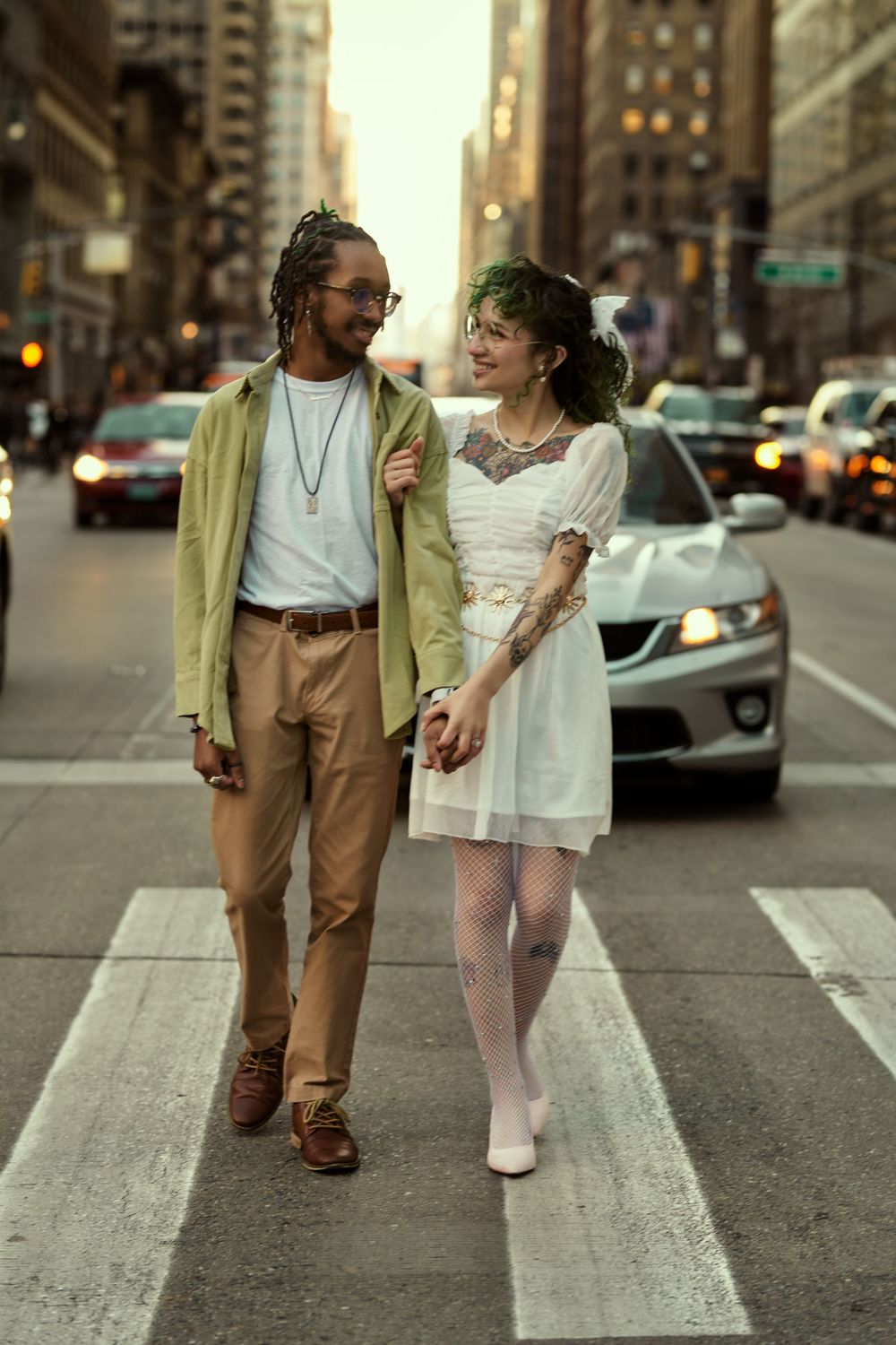 Two individuals in contrasting outfits walking together on a crosswalk in an urban setting during golden hour. Engagement shoot Manhattan
