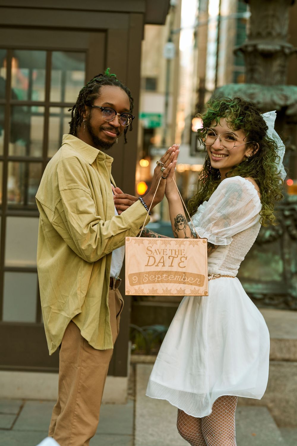A couple in casual attire on a city street sharing a playful moment while holding a wooden Save the Date sign. Engagement shoot Manhattan