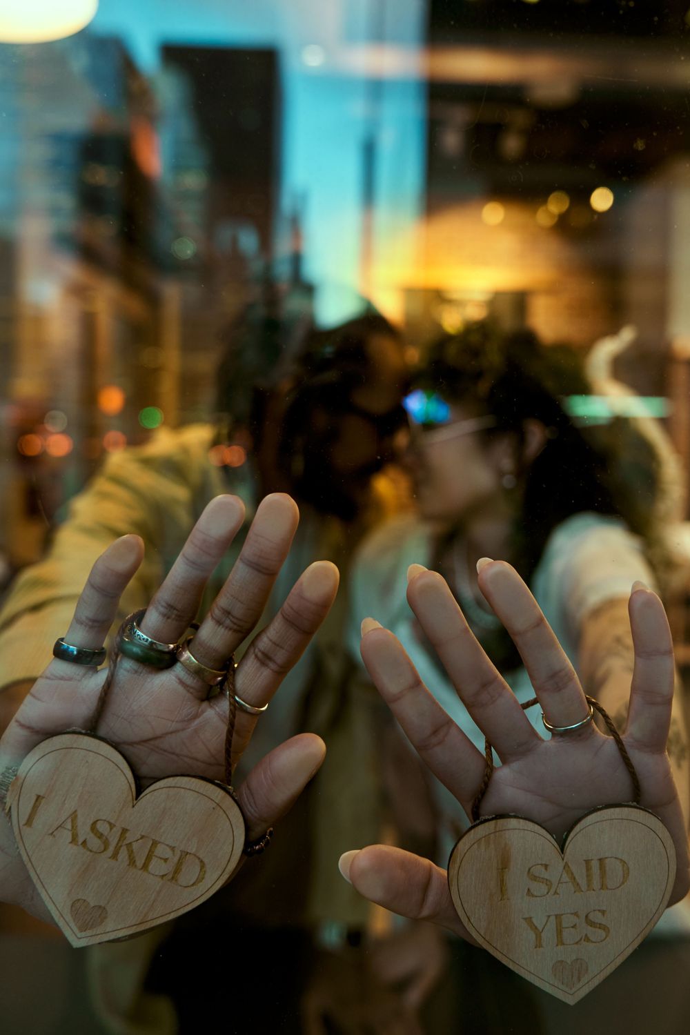 A series of photos showing hands holding up wooden heart signs spelling out 'He asked I said yes' in a dimly lit setting. Engagement shoot