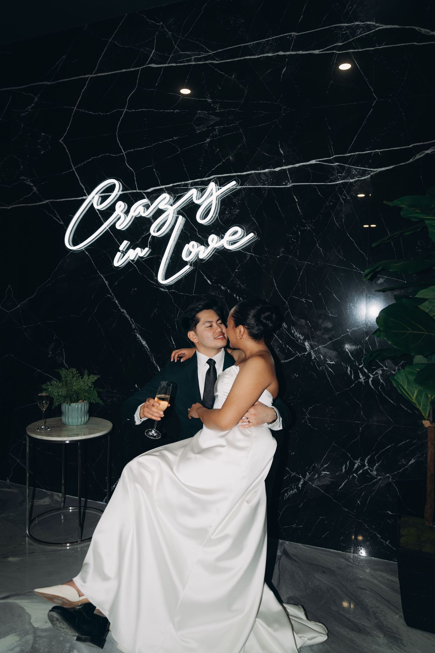 Wedding couple embraces in front of twinkling lights and greenery at an outdoor evening venue.