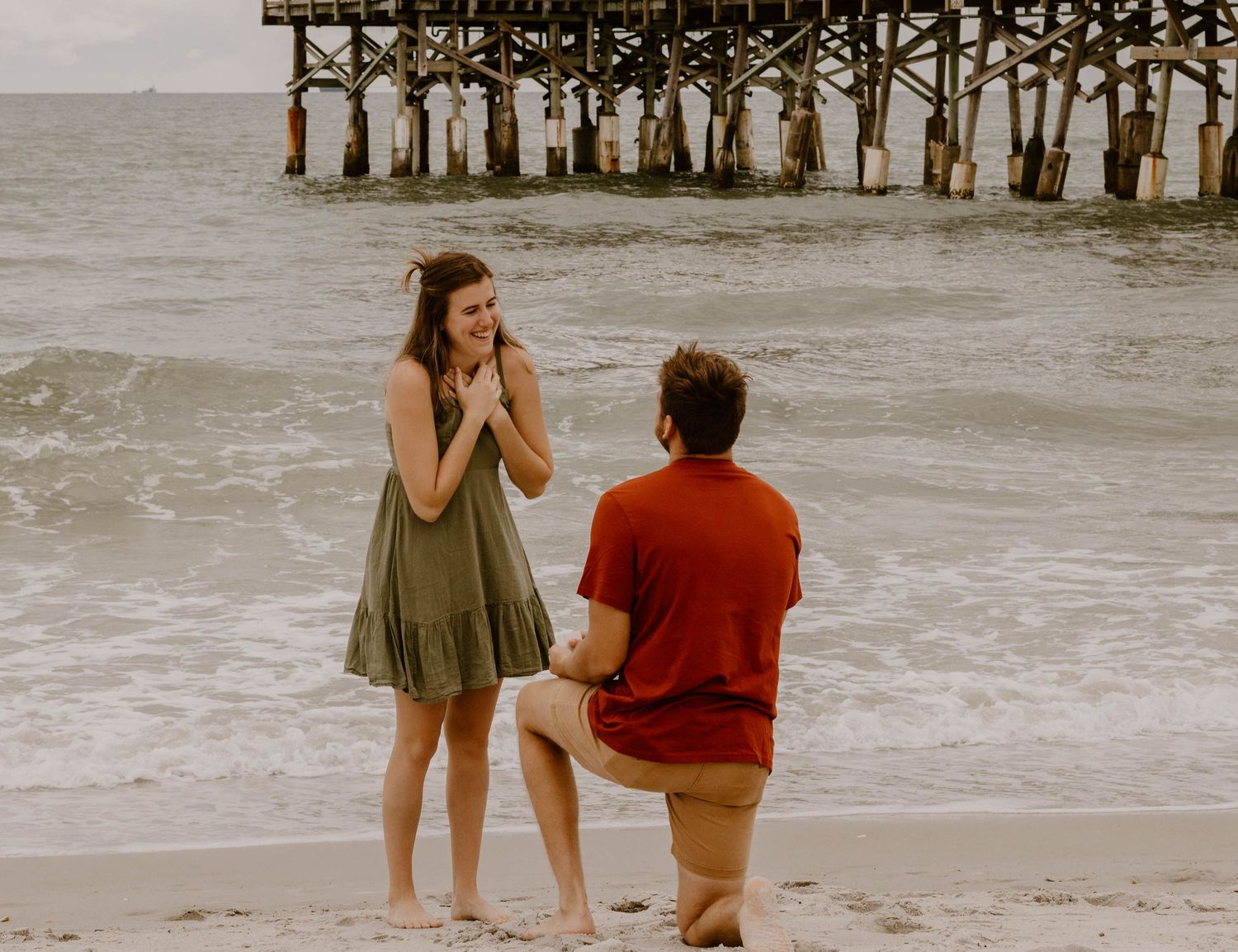 Beach proposal scene with couple by wooden pier during sunset.