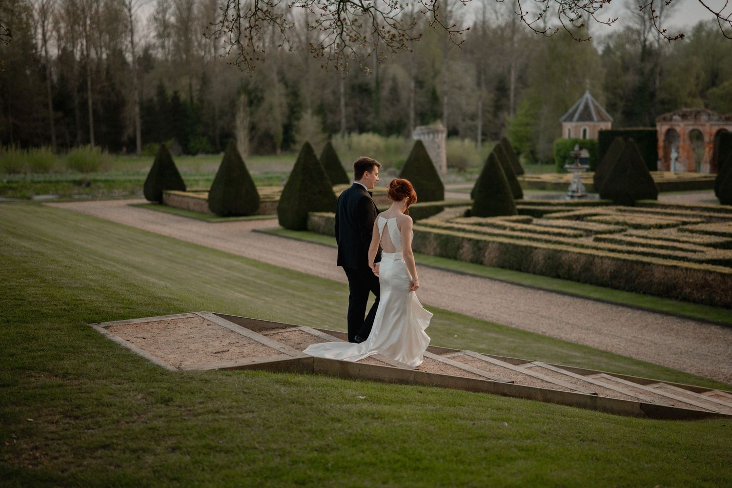 Two people walking through a lush garden landscape with stone pathways and manicured hedges on an overcast day.