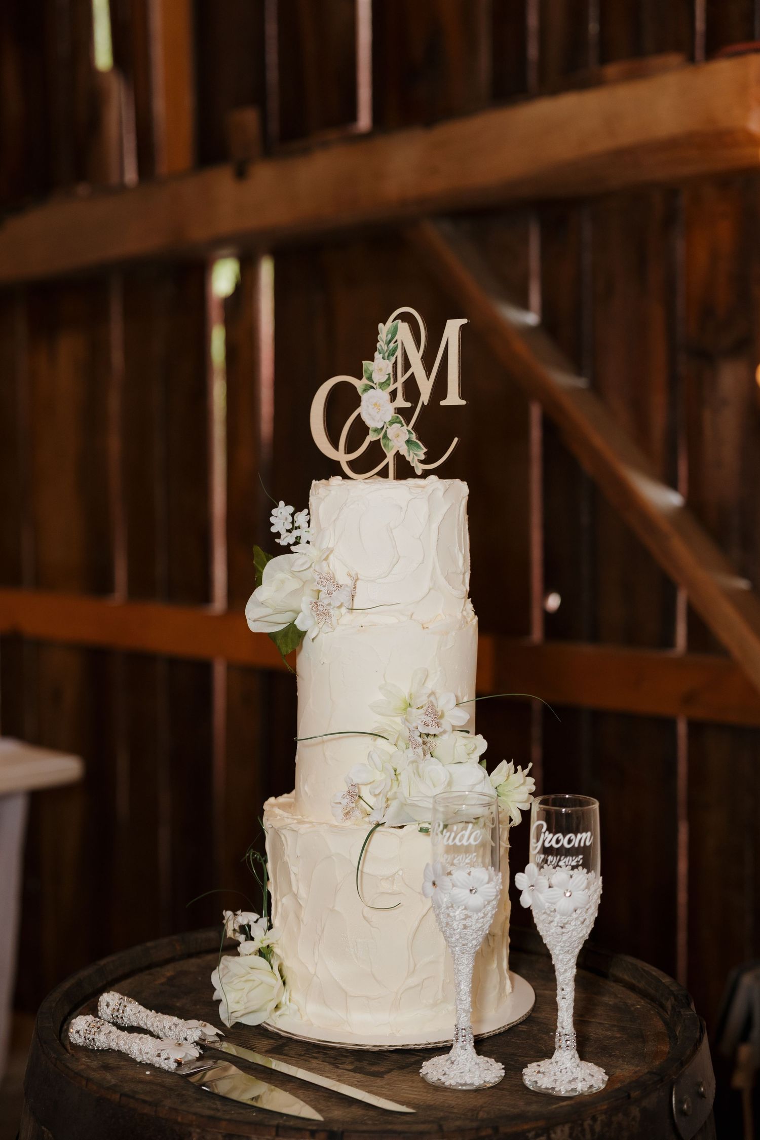 Elegant three-tier white wedding cake with floral decorations and monogram topper displayed next to decorated champagne glasses.