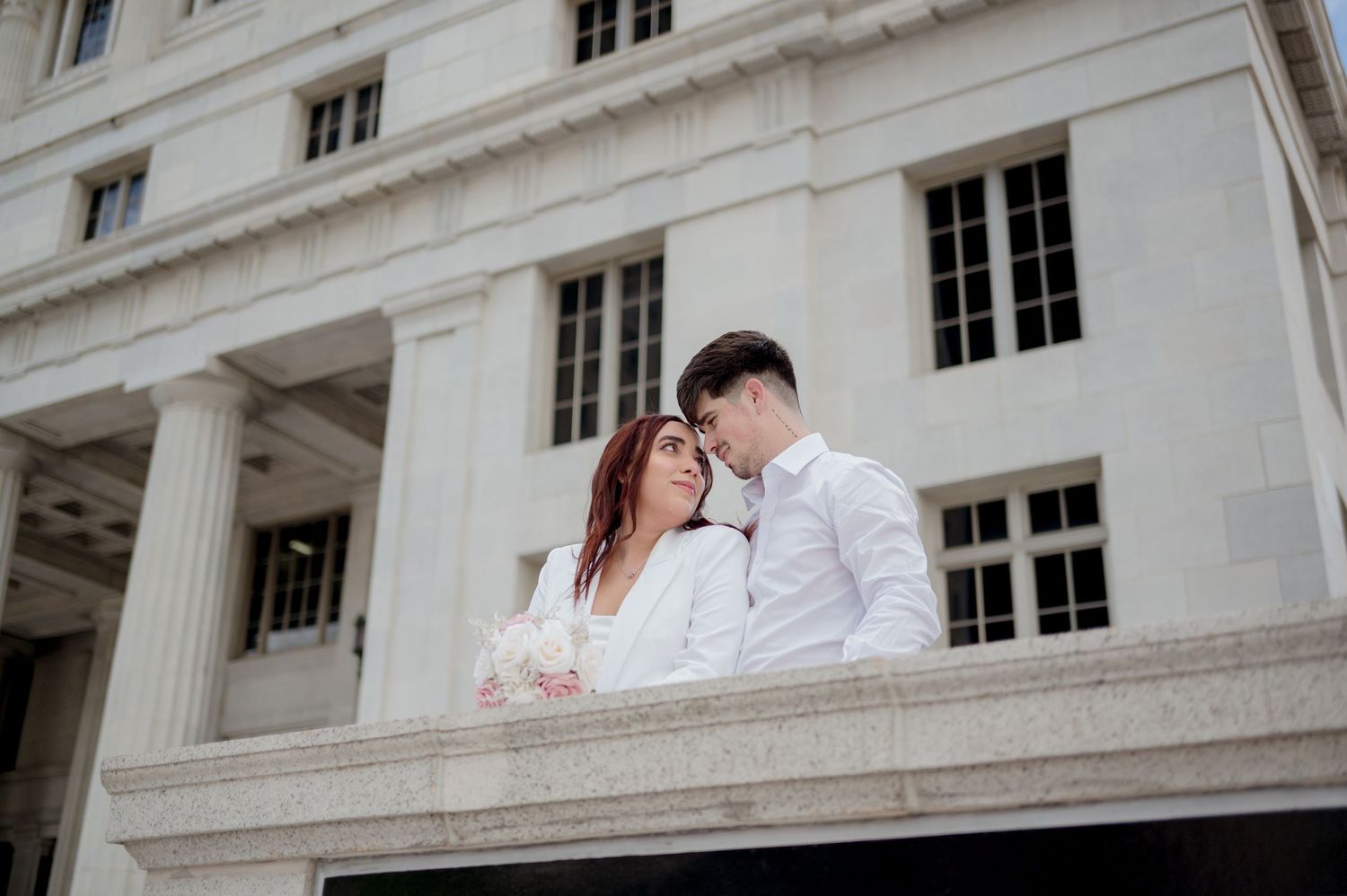 A couple in formal wedding attire poses together on the stairs of a grand white stone building with columns.