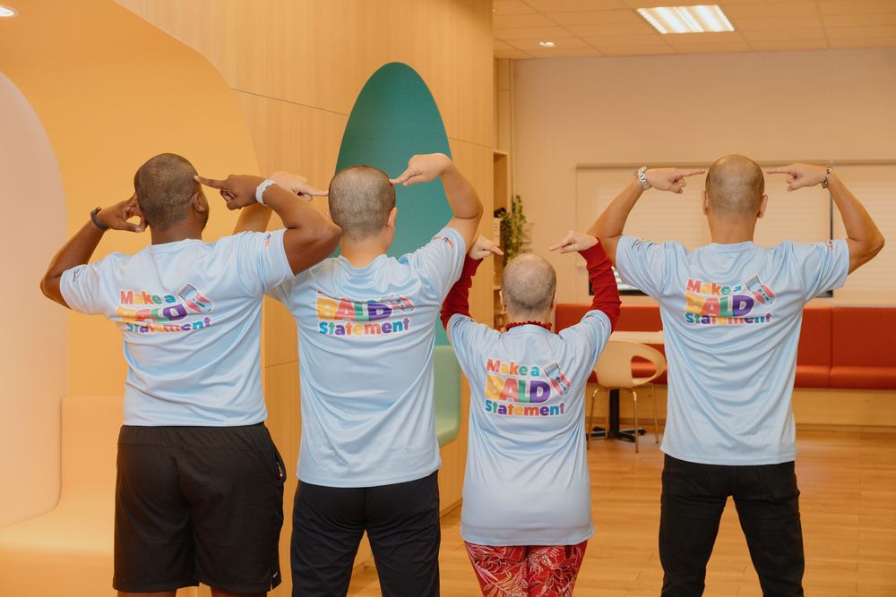 A row of people wearing matching light blue t-shirts stand with their backs facing the camera in a large indoor space.