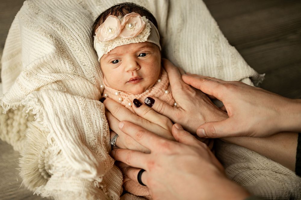 Newborn baby girl in cream wrap with floral headband, cradled gently by parents hands in a soft studio portrait.