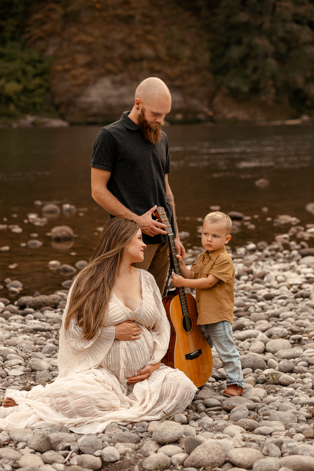 Pregnant woman in white dress sits on rocky riverbank with bearded man and toddler holding acoustic guitar.