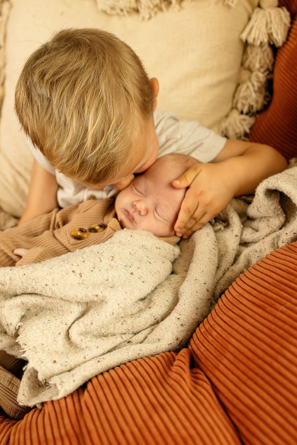 Older blonde boy gently kissing sleeping newborn baby wrapped in cozy knit blanket on warm cushioned sofa.