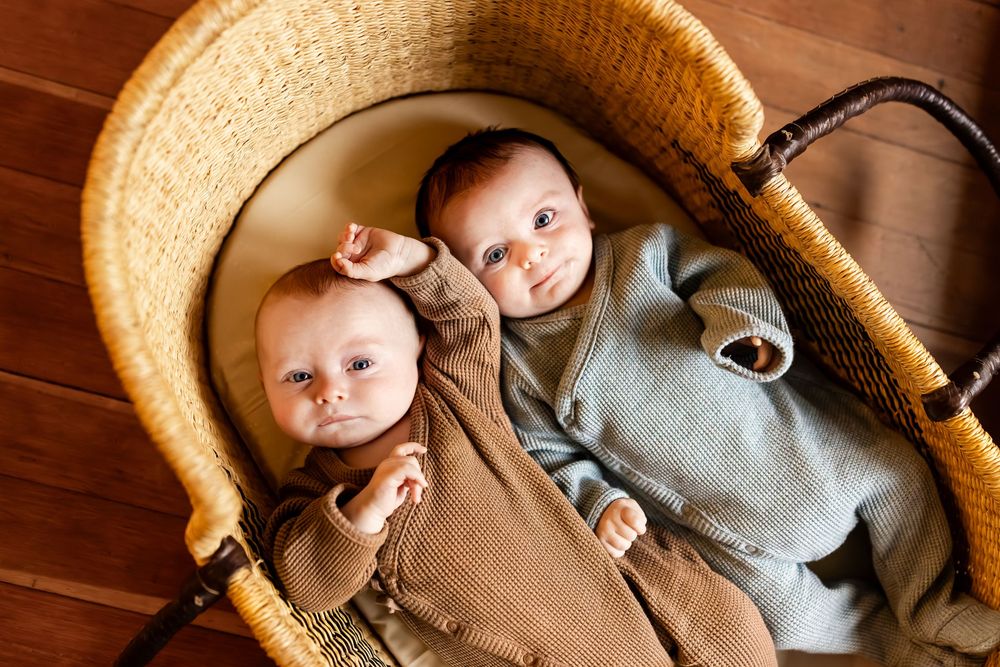 Two newborn twins lying together in a wicker Moses basket, wearing brown and blue outfits on hardwood floor.
