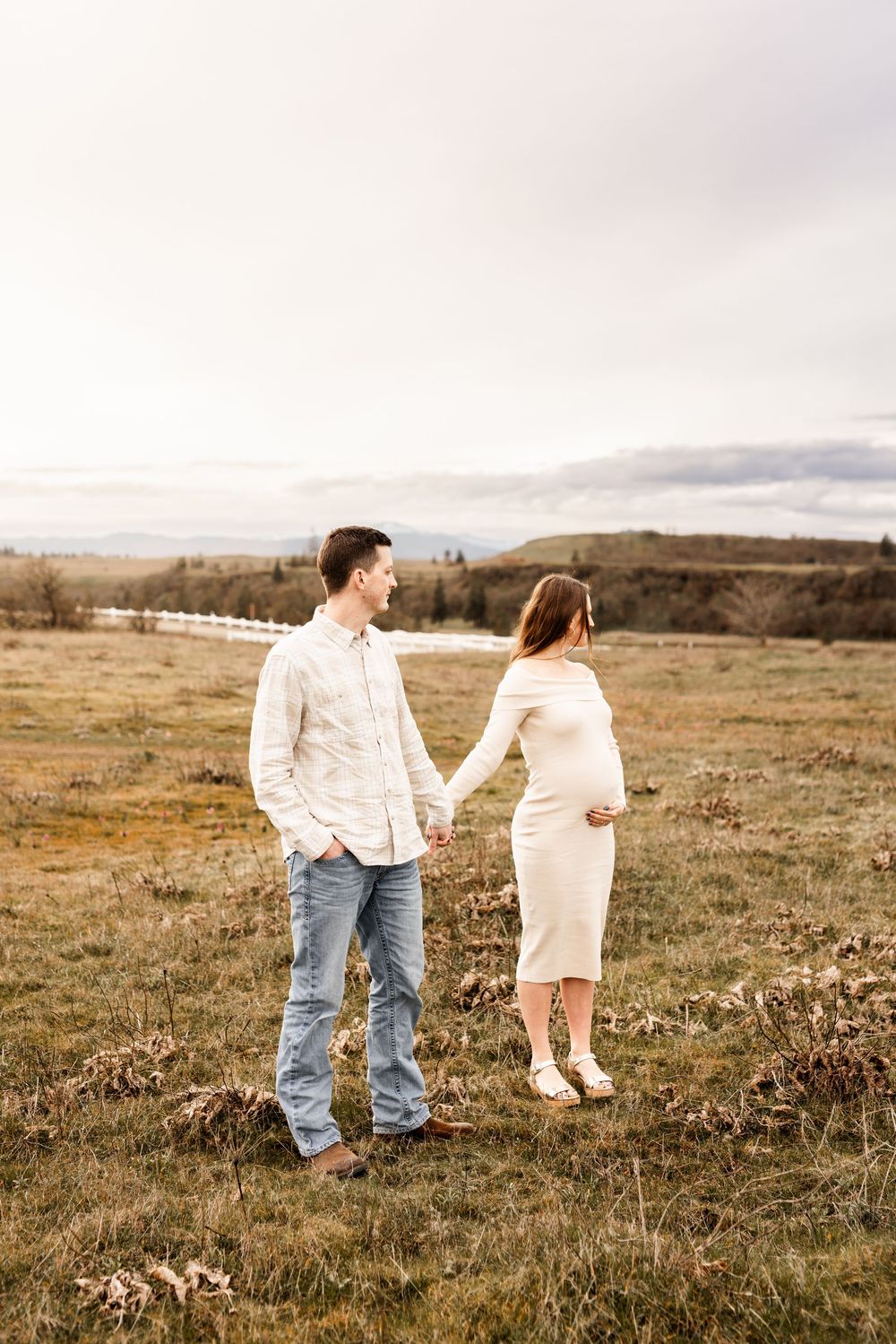 Couple holding hands in a field, woman in white maternity dress, man in jeans and white shirt, rural landscape backdrop.