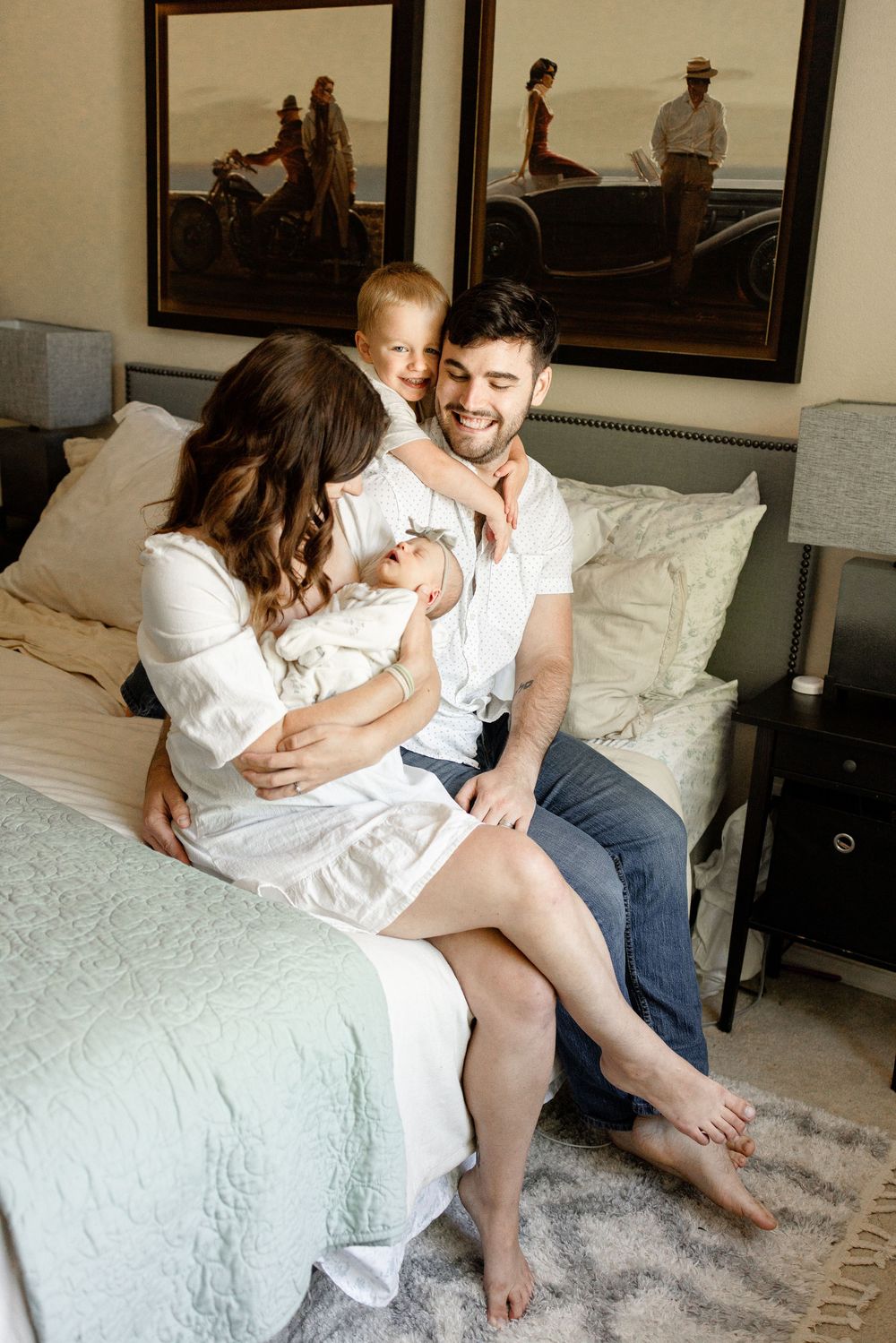 Happy family of four sitting on a bed, parents holding newborn and toddler, smiling in a cozy bedroom.