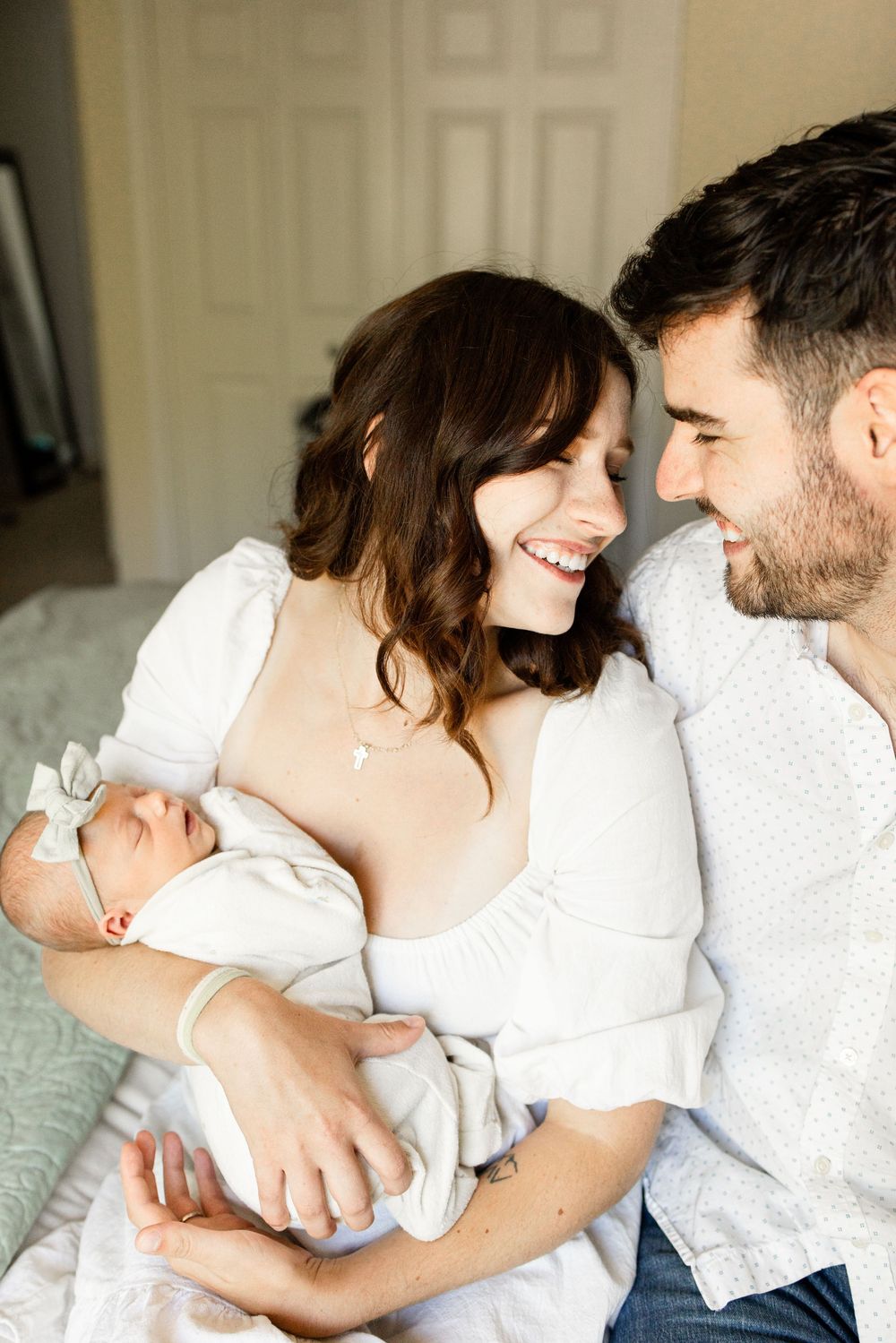 Happy new parents smiling at each other while cradling their sleeping newborn baby wrapped in white.
