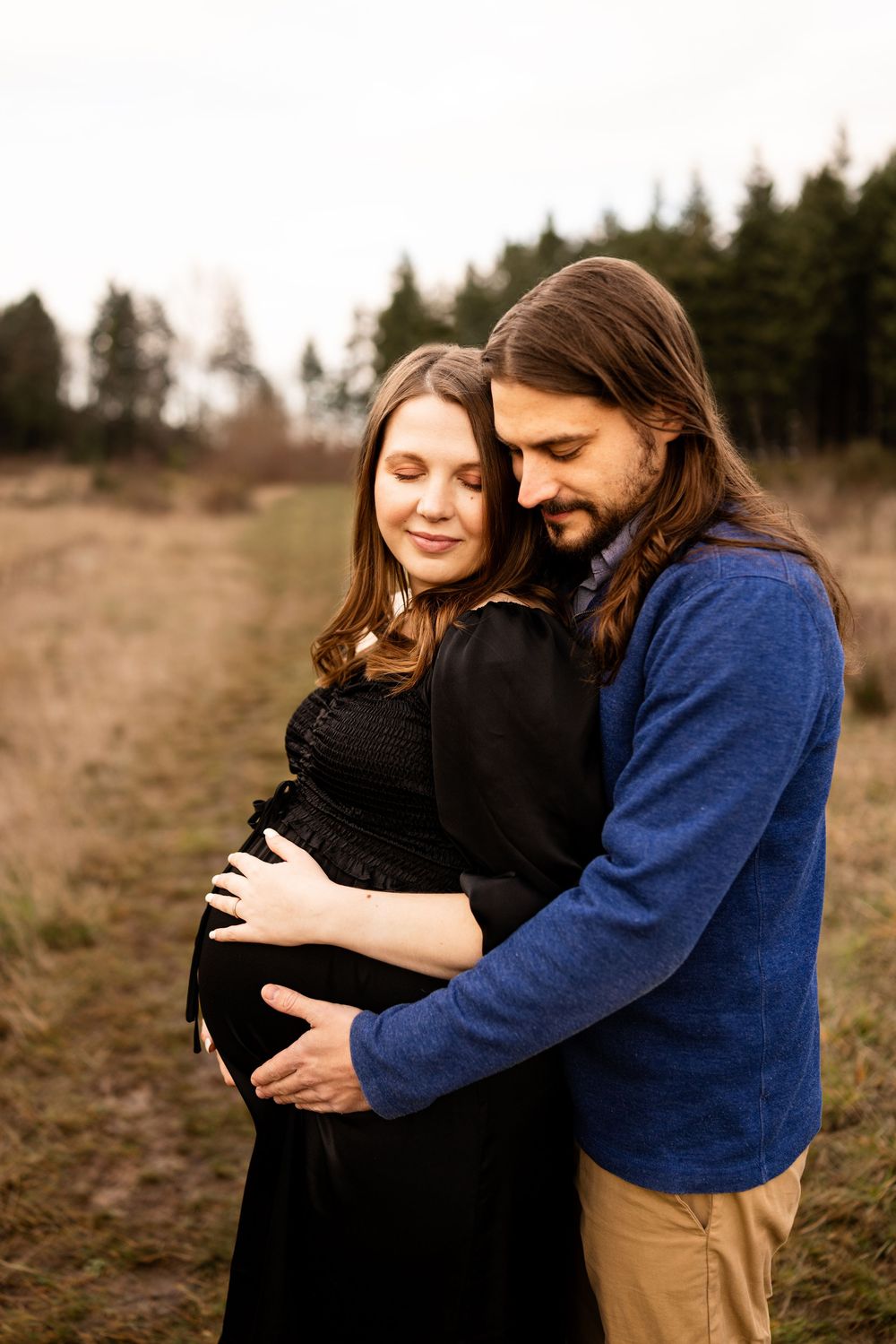 A couple tenderly embraces outdoors, the pregnant woman in black dress, partner in blue sweater, hands on her baby bump.