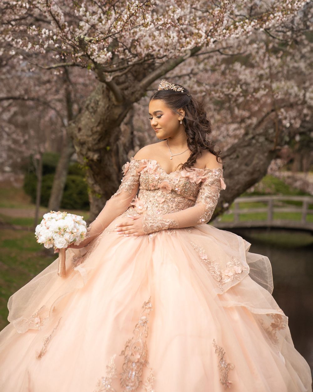 A person in a blush pink ball gown with floral lace details poses under blooming cherry blossom trees holding a white bouquet.