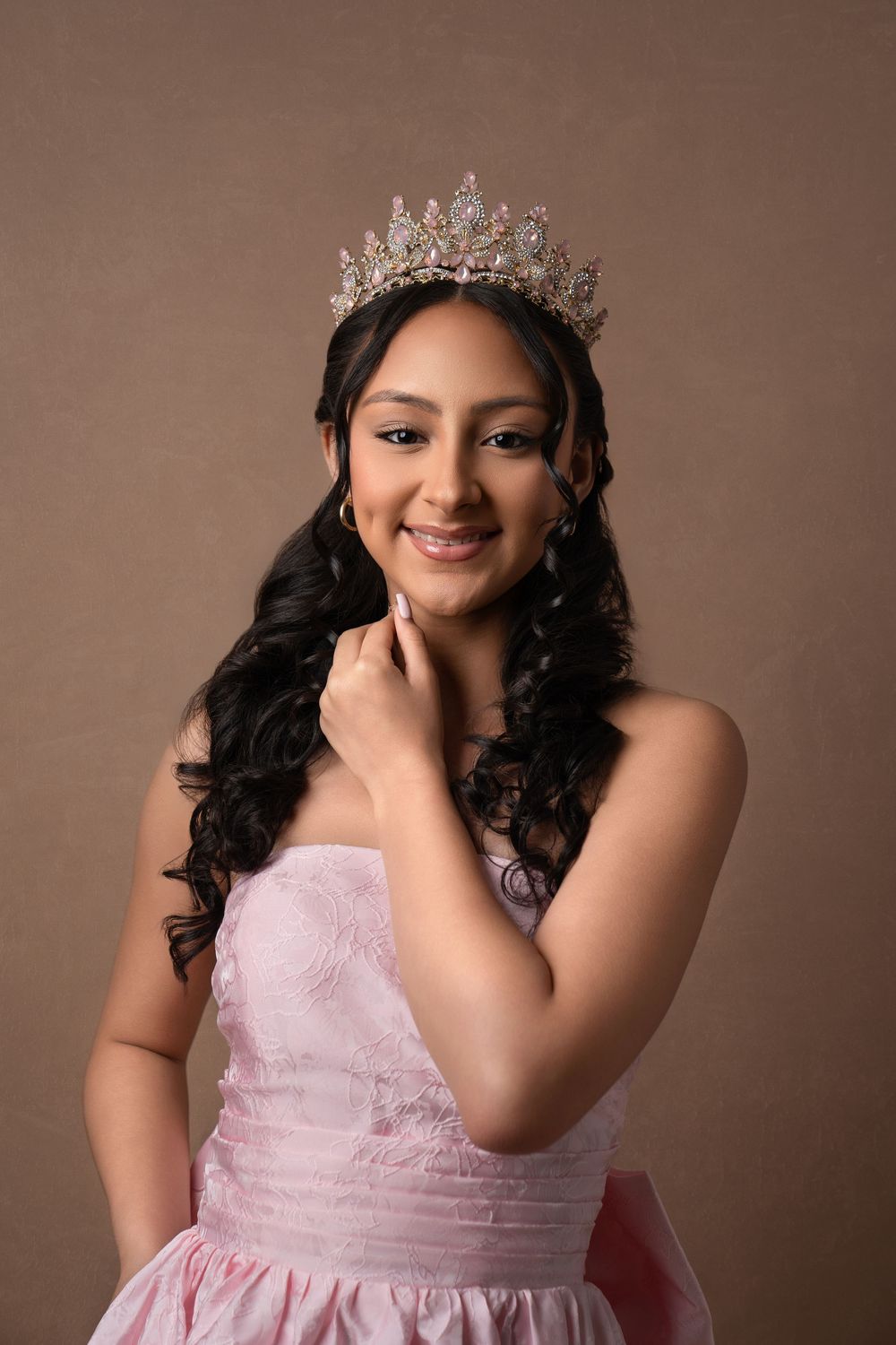 Portrait of someone wearing a crystal crown and pink strapless dress with soft curled dark hair against brown backdrop.