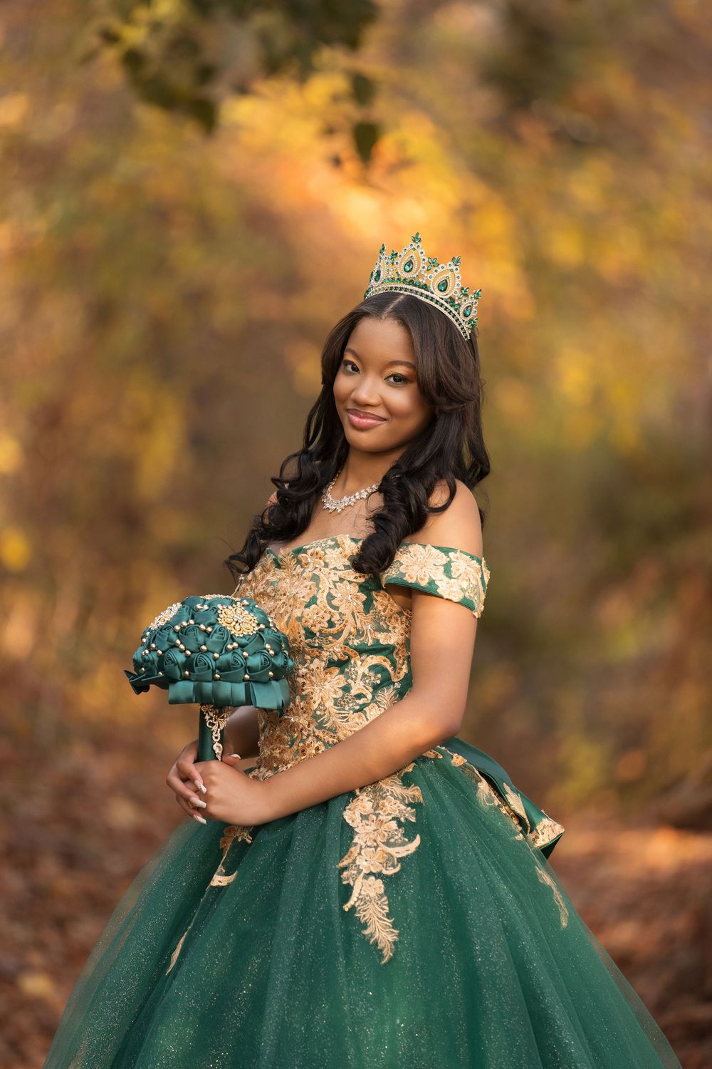 Elegant formal portrait showing a sparkling emerald green ball gown with gold accents and a crown against autumn foliage.
